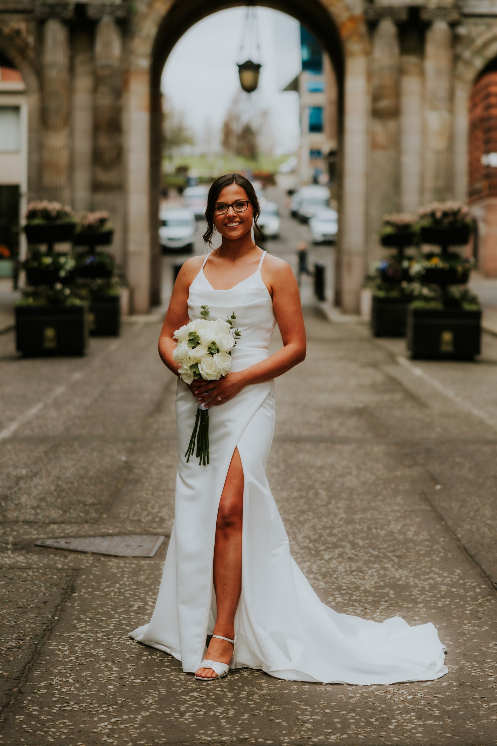 Alana & Kyle’s Glasgow Wedding — City Centre to The Bothy 15 Bride in sleeveless white dress and glasses holding white-rose bouquet under a stone city-centre arch in Glasgow.