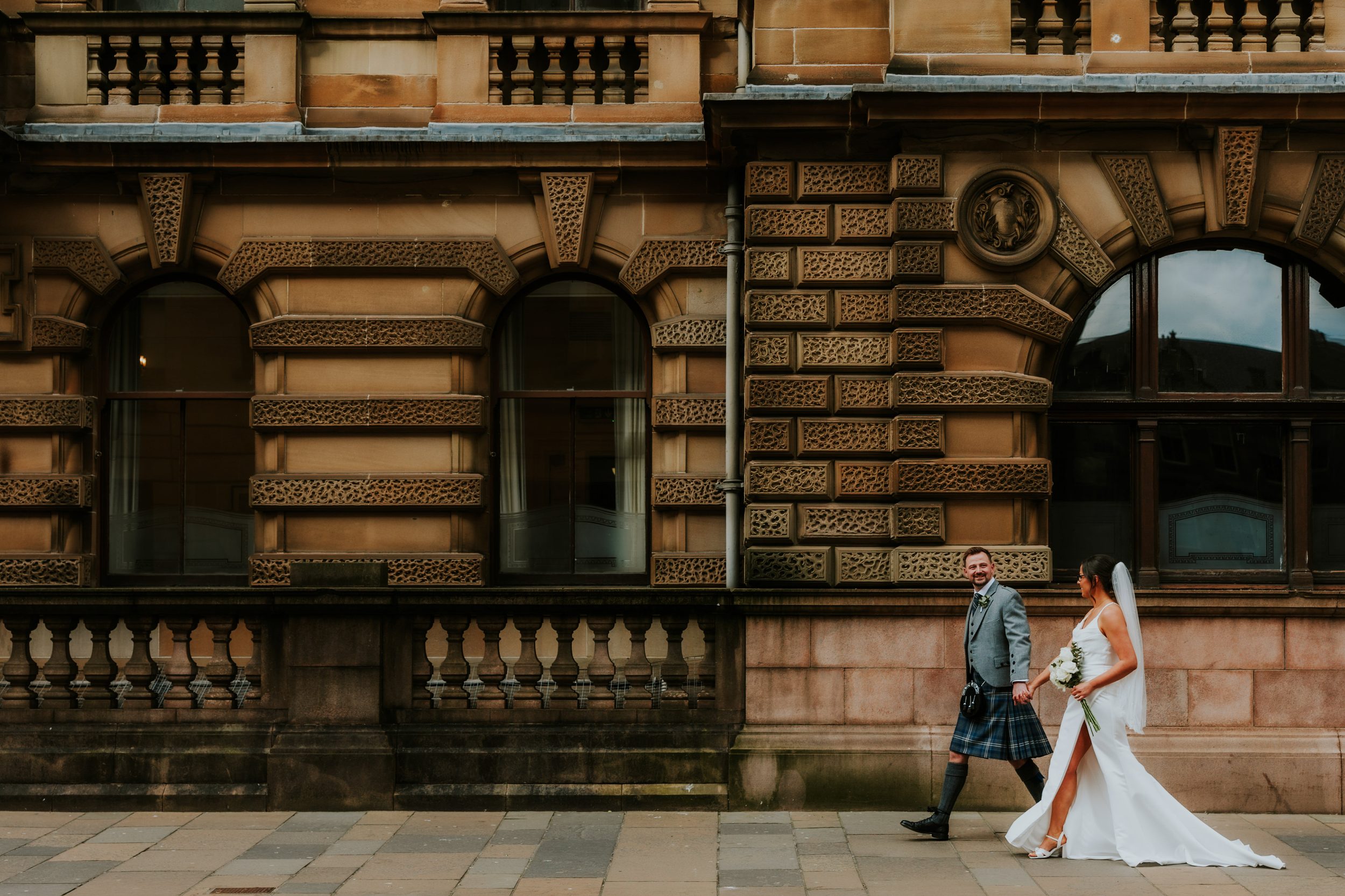 Alana & Kyle’s Glasgow Wedding — City Centre to The Bothy 13 Bride in white dress and groom in kilt walking past an ornate sandstone building in Glasgow.