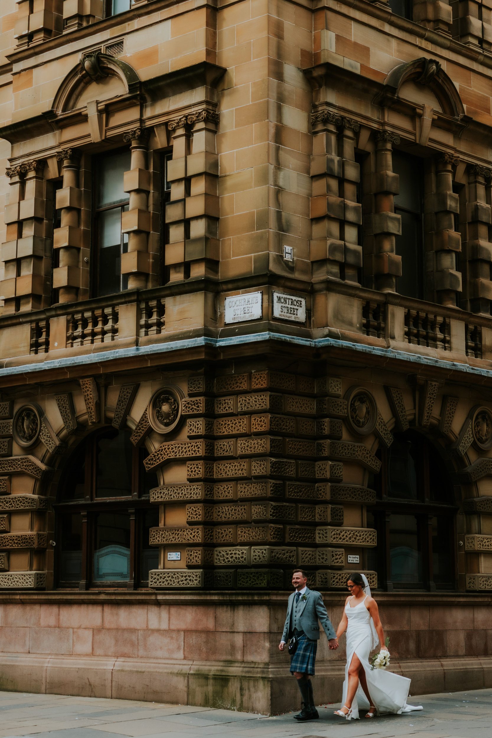 Alana & Kyle’s Glasgow Wedding — City Centre to The Bothy 20 Bride in white dress and groom in kilt holding hands walking past a carved sandstone city-centre building in Glasgow.