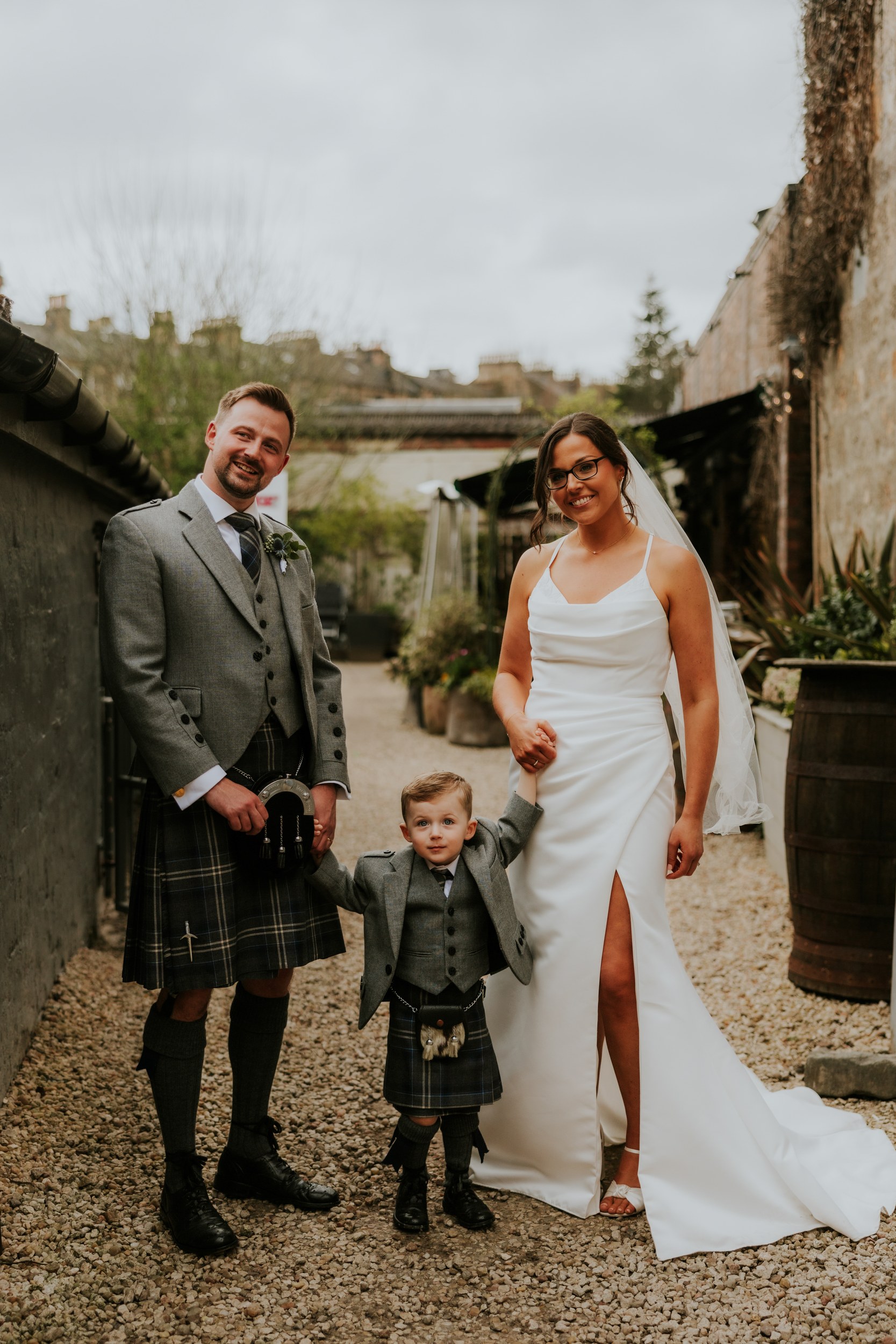 Alana & Kyle’s Glasgow Wedding — City Centre to The Bothy 26 Bride in white dress and groom in kilt holding a young boy in a gravel courtyard at The Bothy.