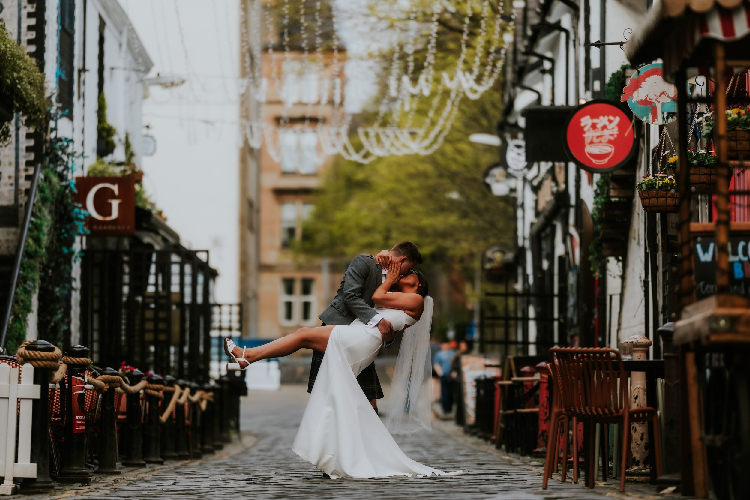 Alana & Kyle’s Glasgow Wedding — City Centre to The Bothy 29 Bride in white dress and veil dipped and kissed by groom in kilt on a cobbled Glasgow lane with hanging lights and outdoor seating.
