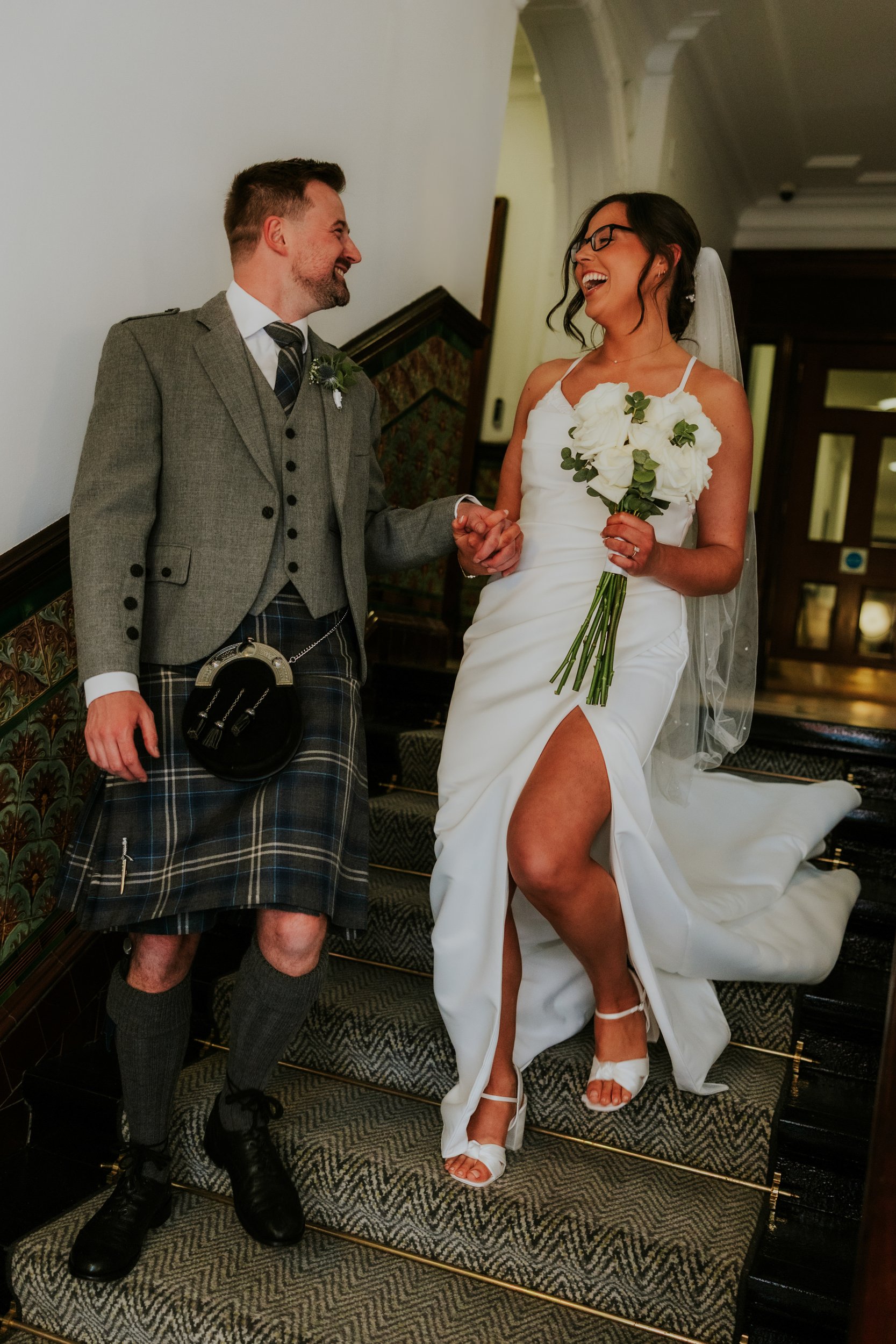 Alana & Kyle’s Glasgow Wedding — City Centre to The Bothy 11 Bride in white dress and veil laughing with groom in grey jacket and tartan kilt while holding hands on a staircase, bride holds white-rose bouquet.