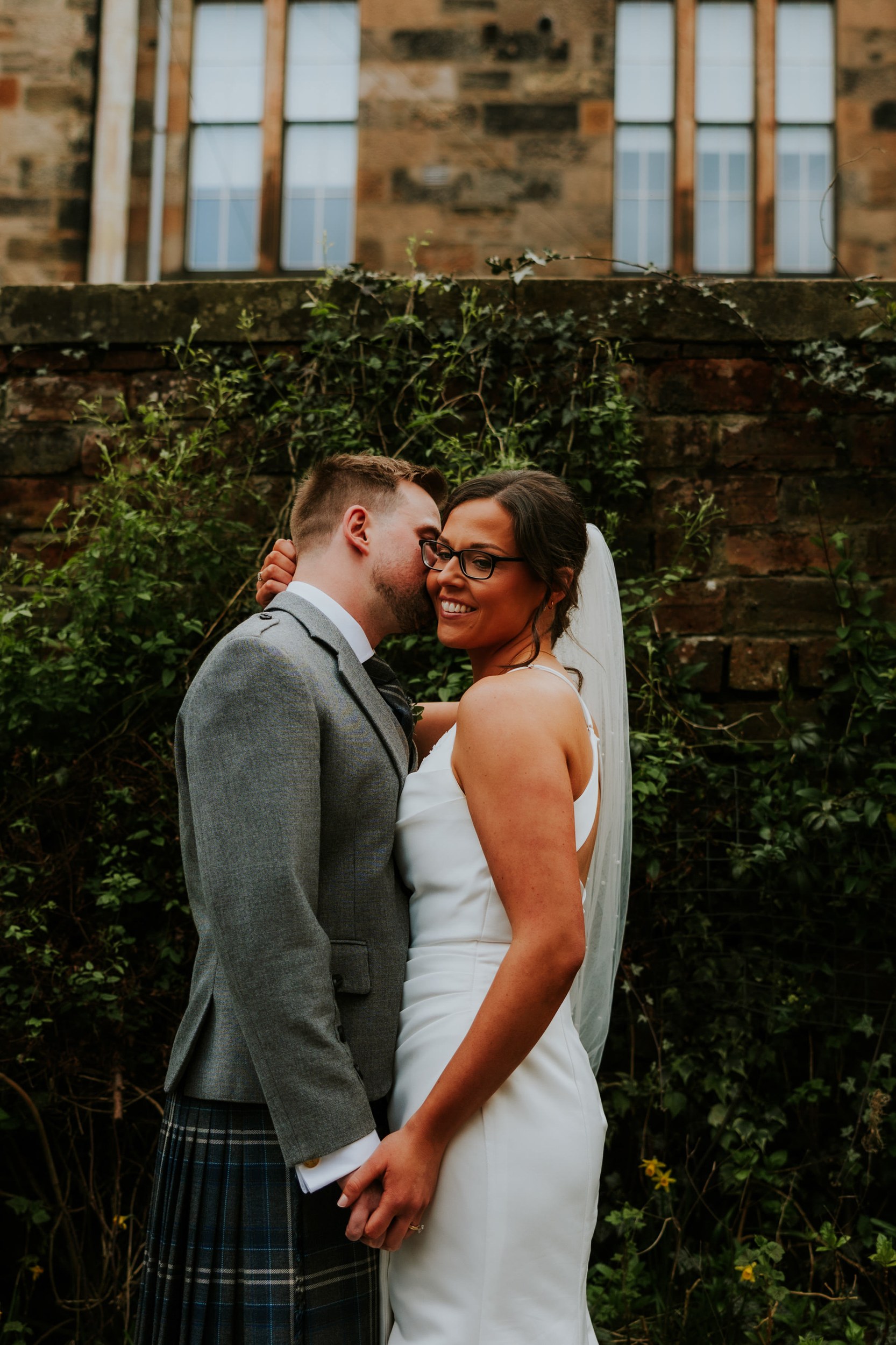 Alana & Kyle’s Glasgow Wedding — City Centre to The Bothy 27 Bride in white dress and veil smiles as groom in kilt kisses her cheek; they hold hands in front of an ivy-covered brick wall.