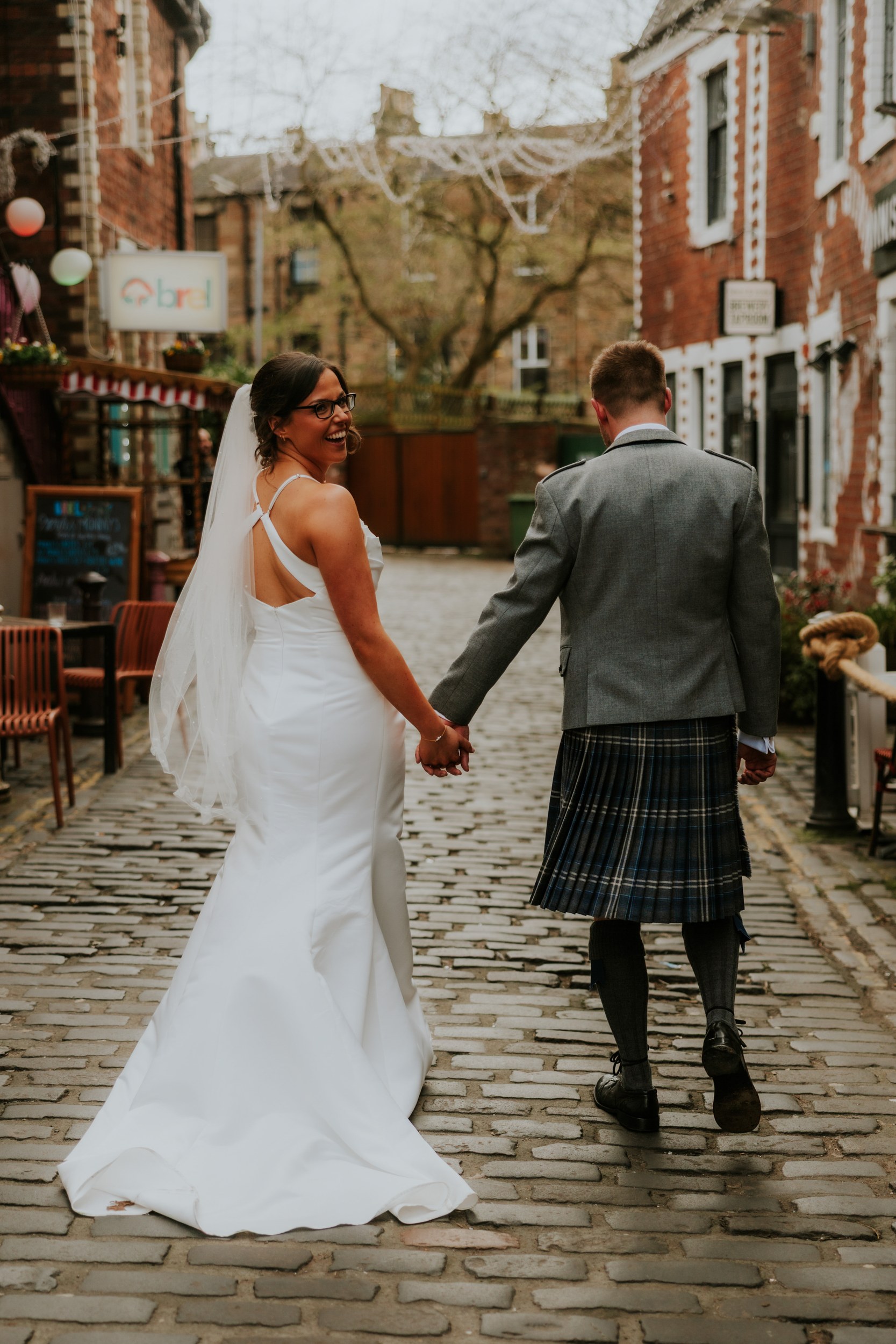 Alana & Kyle’s Glasgow Wedding — City Centre to The Bothy 28 Bride in white dress and veil smiling back while holding hands with groom in kilt on a cobbled street outside The Bothy.
