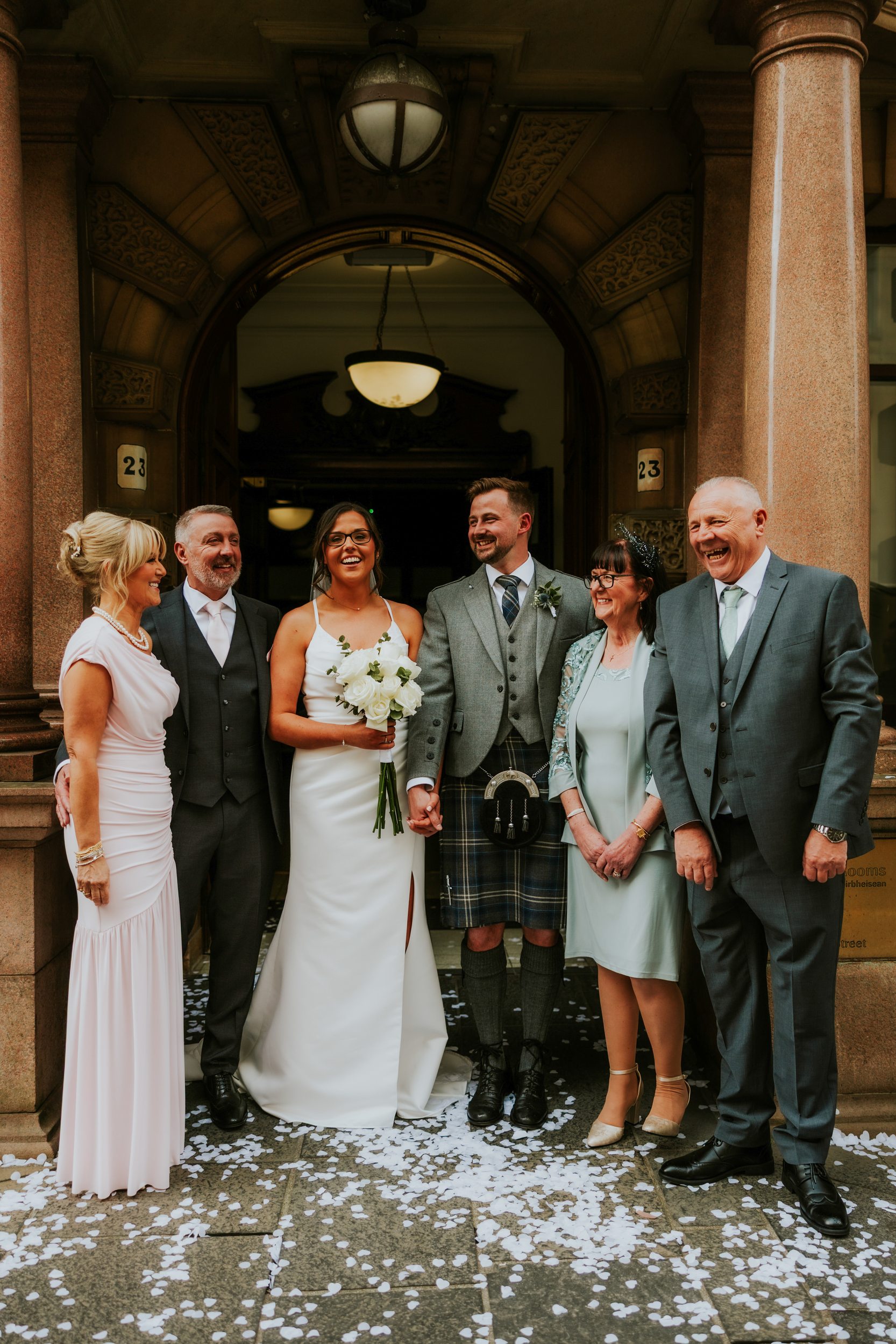 Alana & Kyle’s Glasgow Wedding — City Centre to The Bothy 10 Bride in white dress with bouquet and groom in a kilt smiling with family under an arched stone doorway, confetti on the ground.