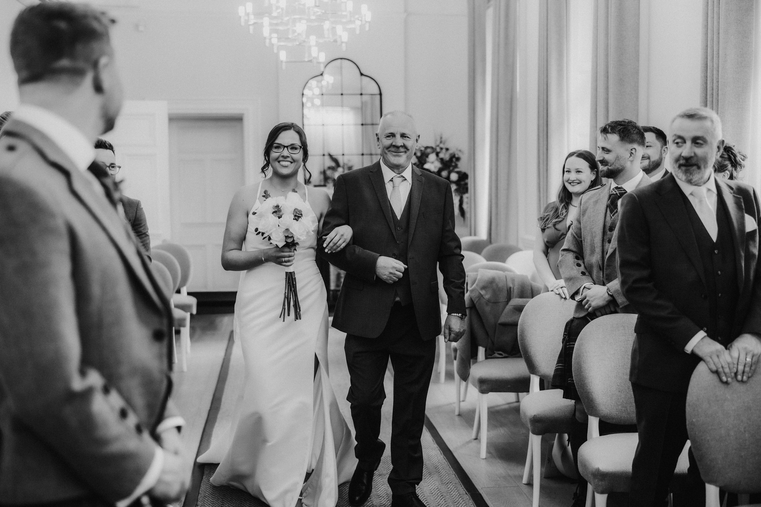Alana & Kyle’s Glasgow Wedding — City Centre to The Bothy 4 Bride walking down the aisle arm-in-arm with an older man as guests and the groom look on in a ceremony room.