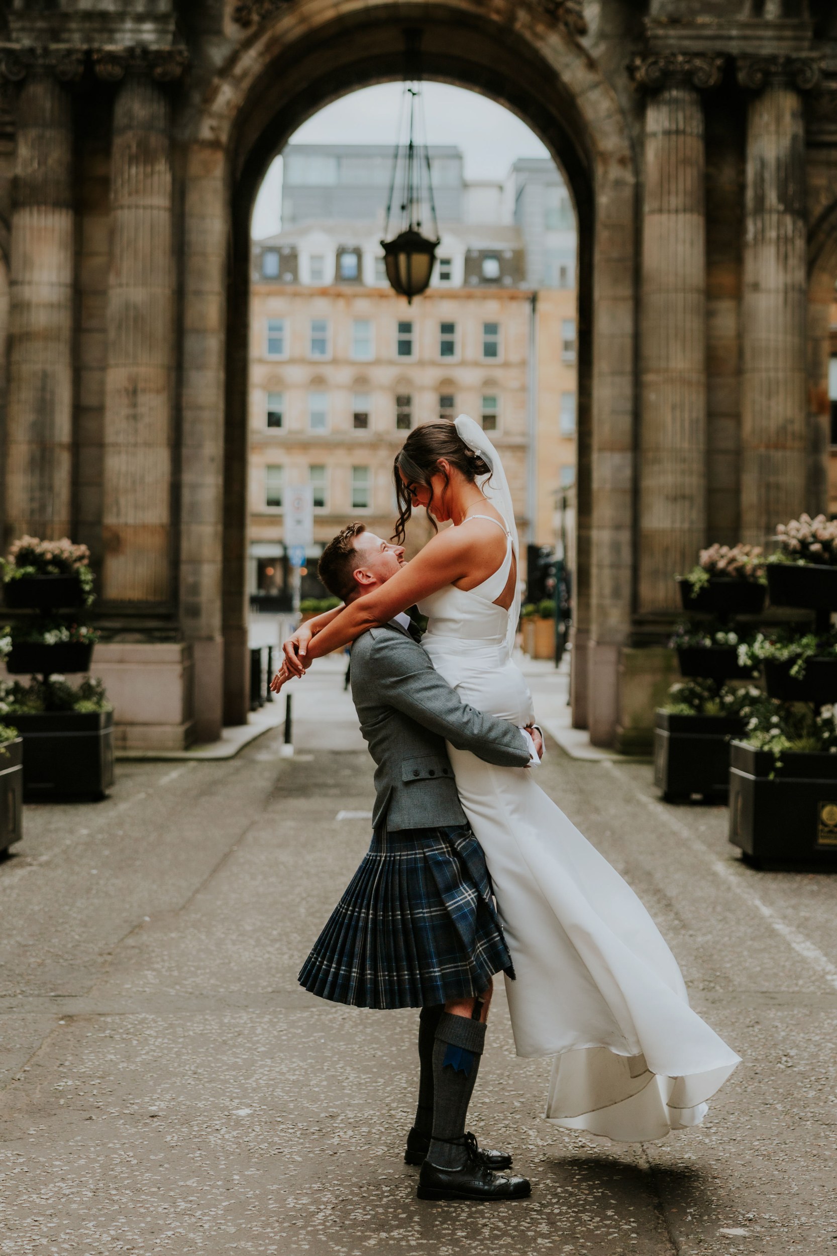 Alana & Kyle’s Glasgow Wedding — City Centre to The Bothy 19 Groom in kilt lifts bride in white dress and veil under a stone arch and hanging lamp at Glasgow's City Chambers.