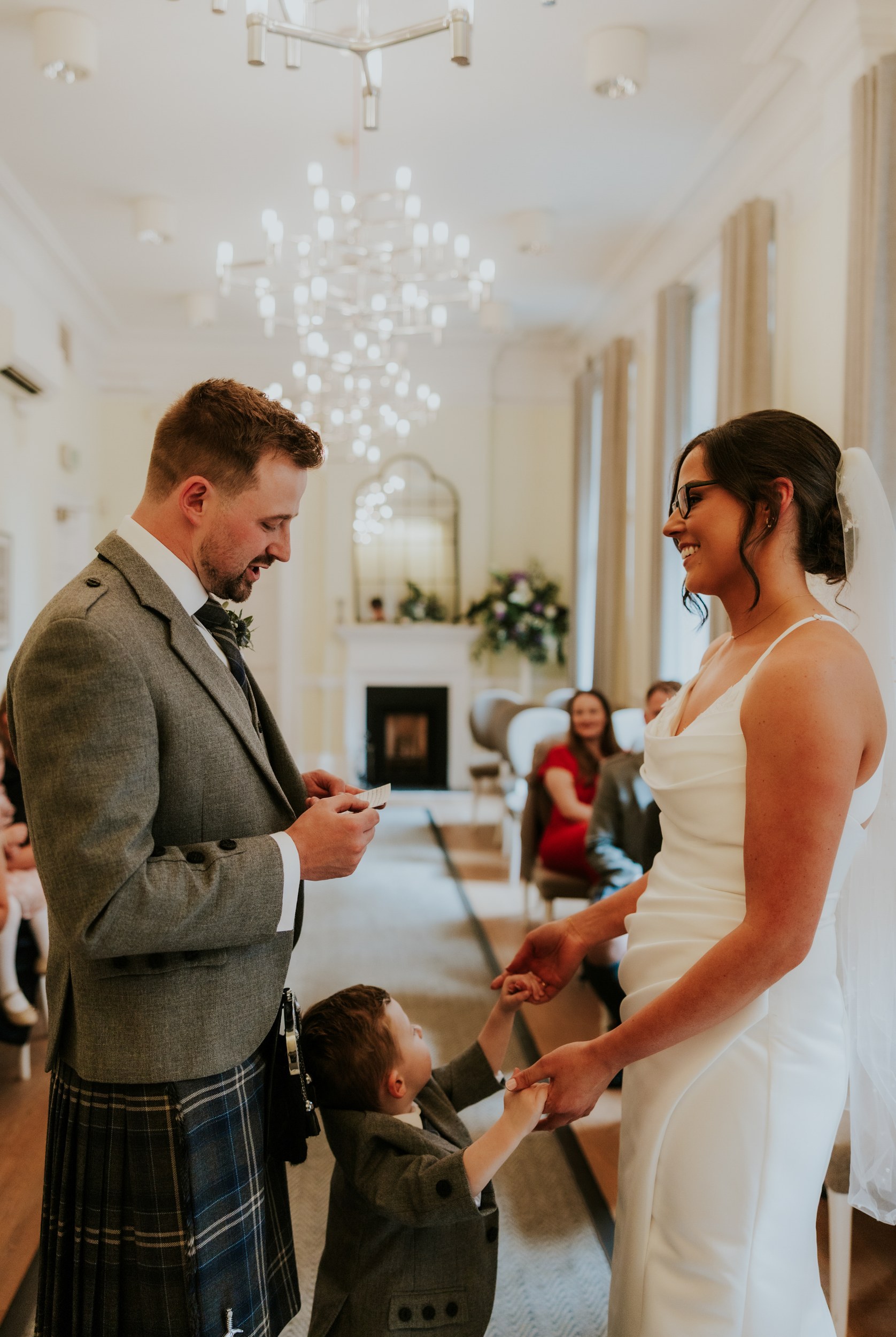 Alana & Kyle’s Glasgow Wedding — City Centre to The Bothy 6 Groom in kilt reads vows as bride smiles and a young boy holds her hands during an indoor ceremony.