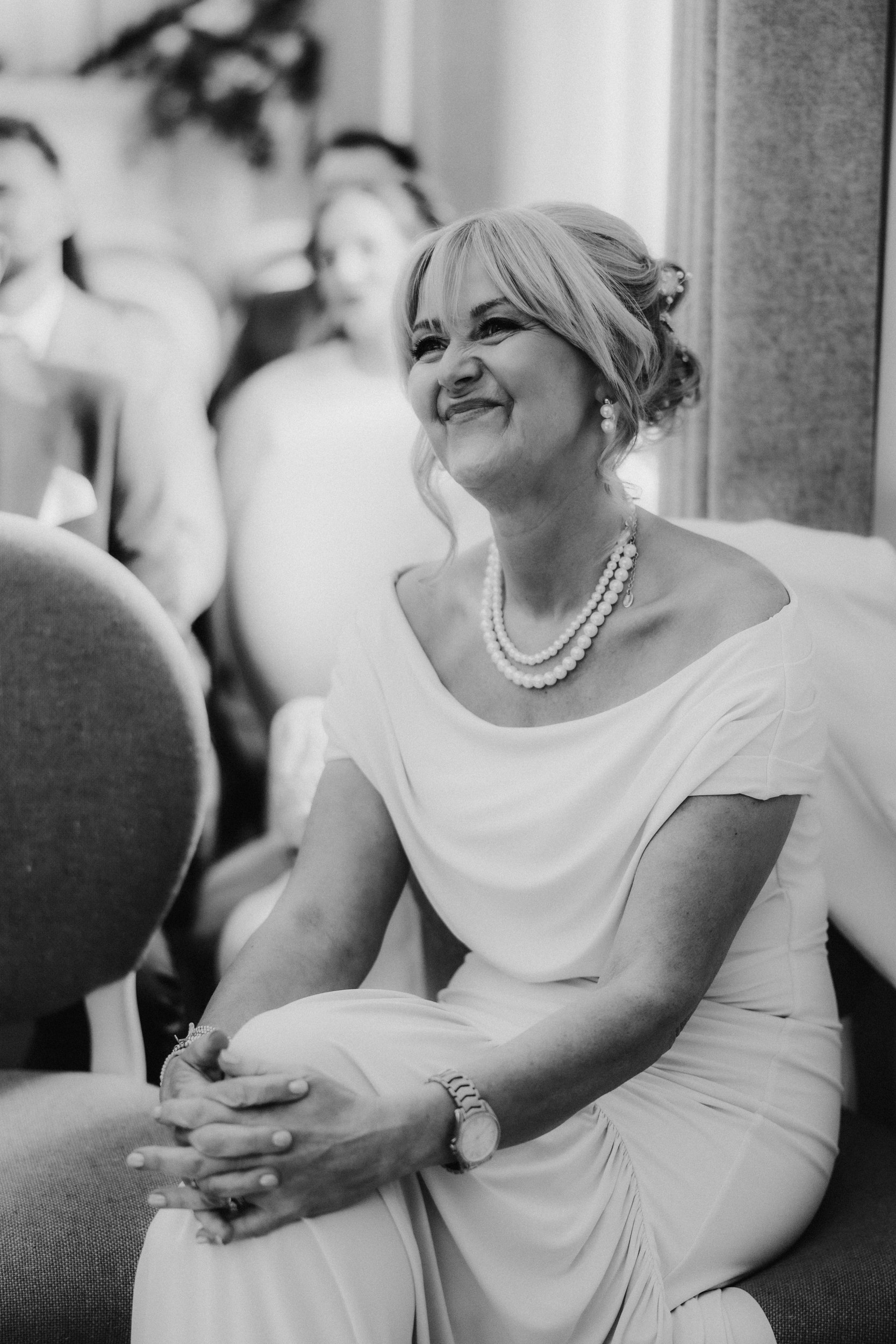 Alana & Kyle’s Glasgow Wedding — City Centre to The Bothy 5 Smiling seated wedding guest in off-shoulder dress wearing layered pearls and a watch, black-and-white image.