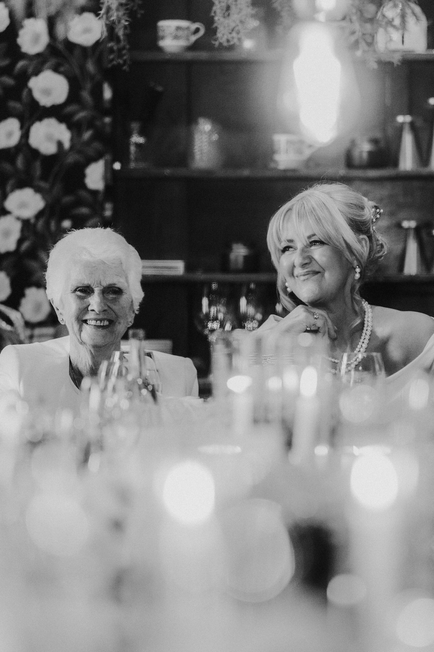 Alana & Kyle’s Glasgow Wedding — City Centre to The Bothy 24 Two women smiling at a wedding table with pearl jewellery, wine glasses and soft bokeh in the foreground.