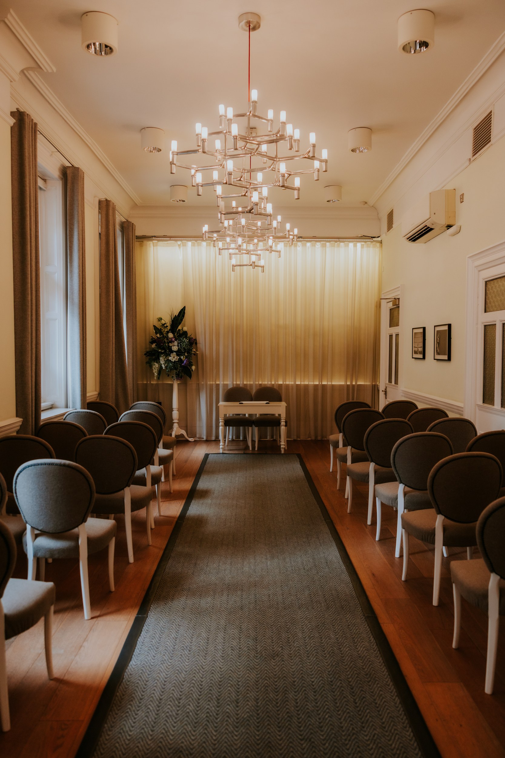 Alana & Kyle’s Glasgow Wedding — City Centre to The Bothy 3 Wedding ceremony room with rows of chairs, central aisle, small signing table and floral arrangement under chandeliers.