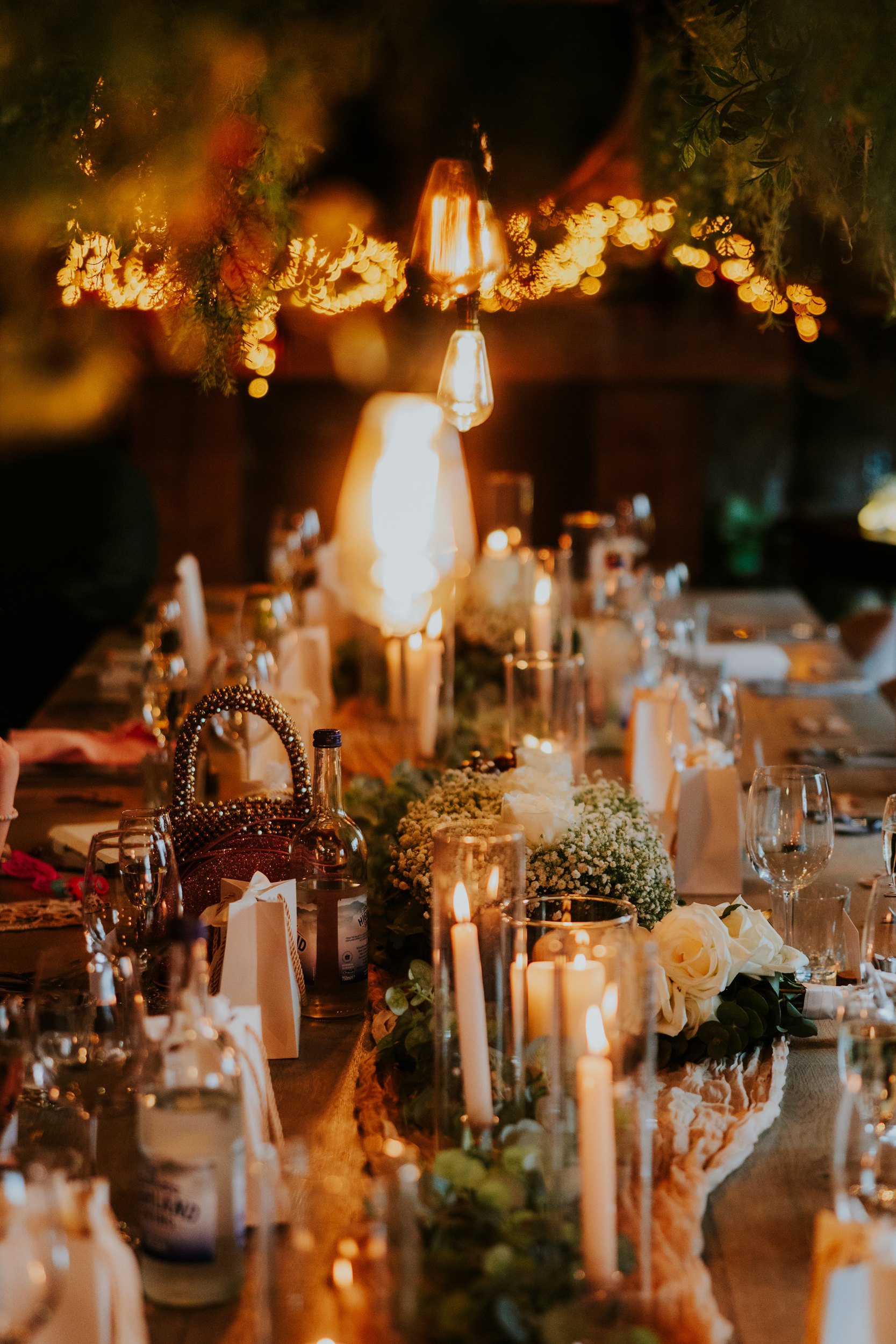 Alana & Kyle’s Glasgow Wedding — City Centre to The Bothy 22 Wedding reception table with candles, glassware, roses and baby's‑breath under hanging filament bulbs and warm fairy lights.