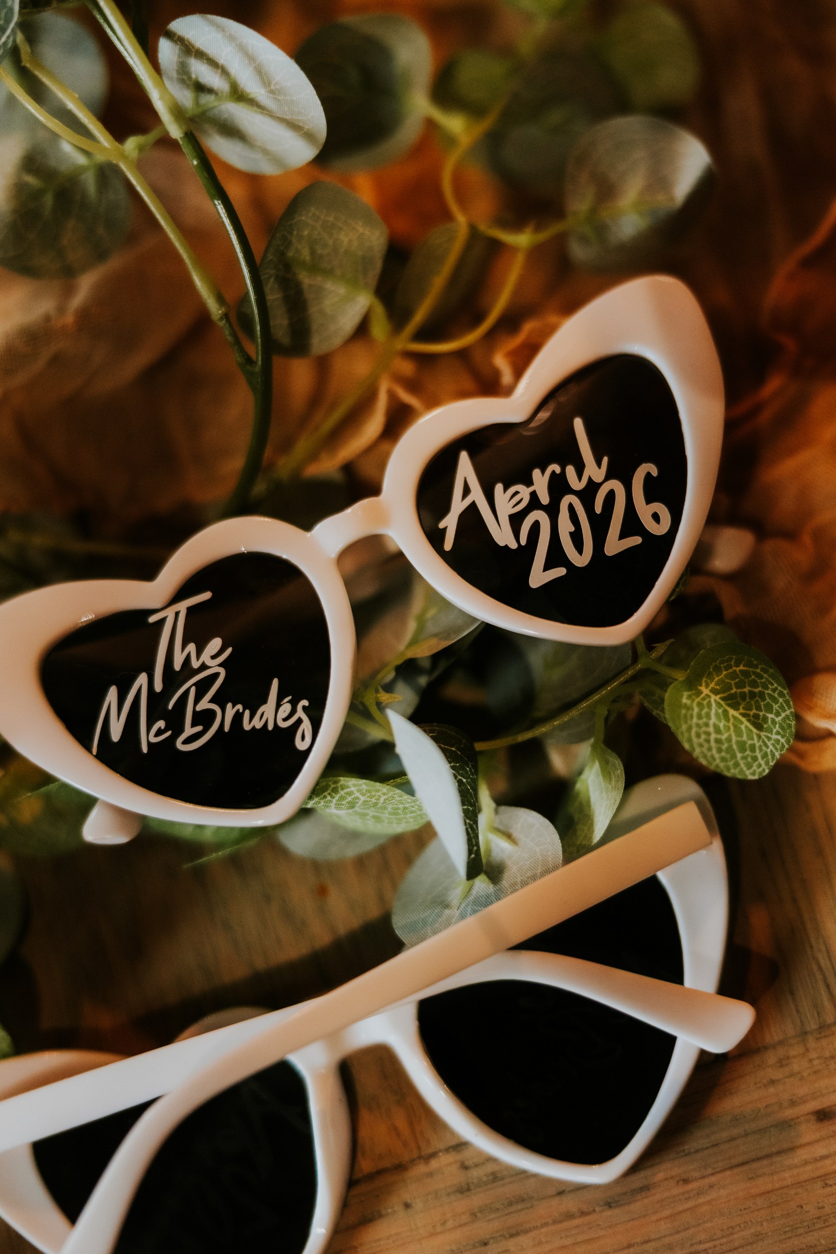 Alana & Kyle’s Glasgow Wedding — City Centre to The Bothy 23 White heart-shaped sunglasses with 'April 2026' written on a lens, placed among eucalyptus leaves on wood.