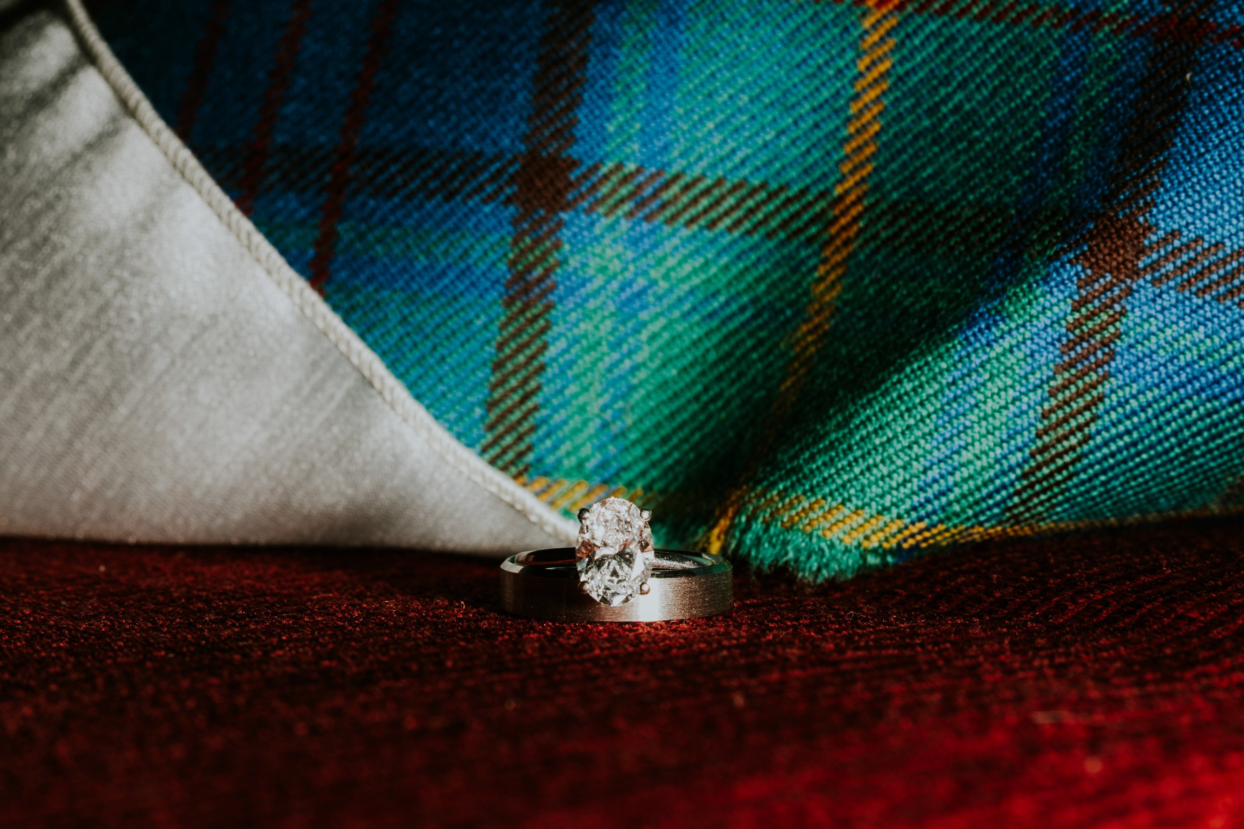 A broad metal band and oval gemstone ring resting on red fabric in front of blue-green tartan.
