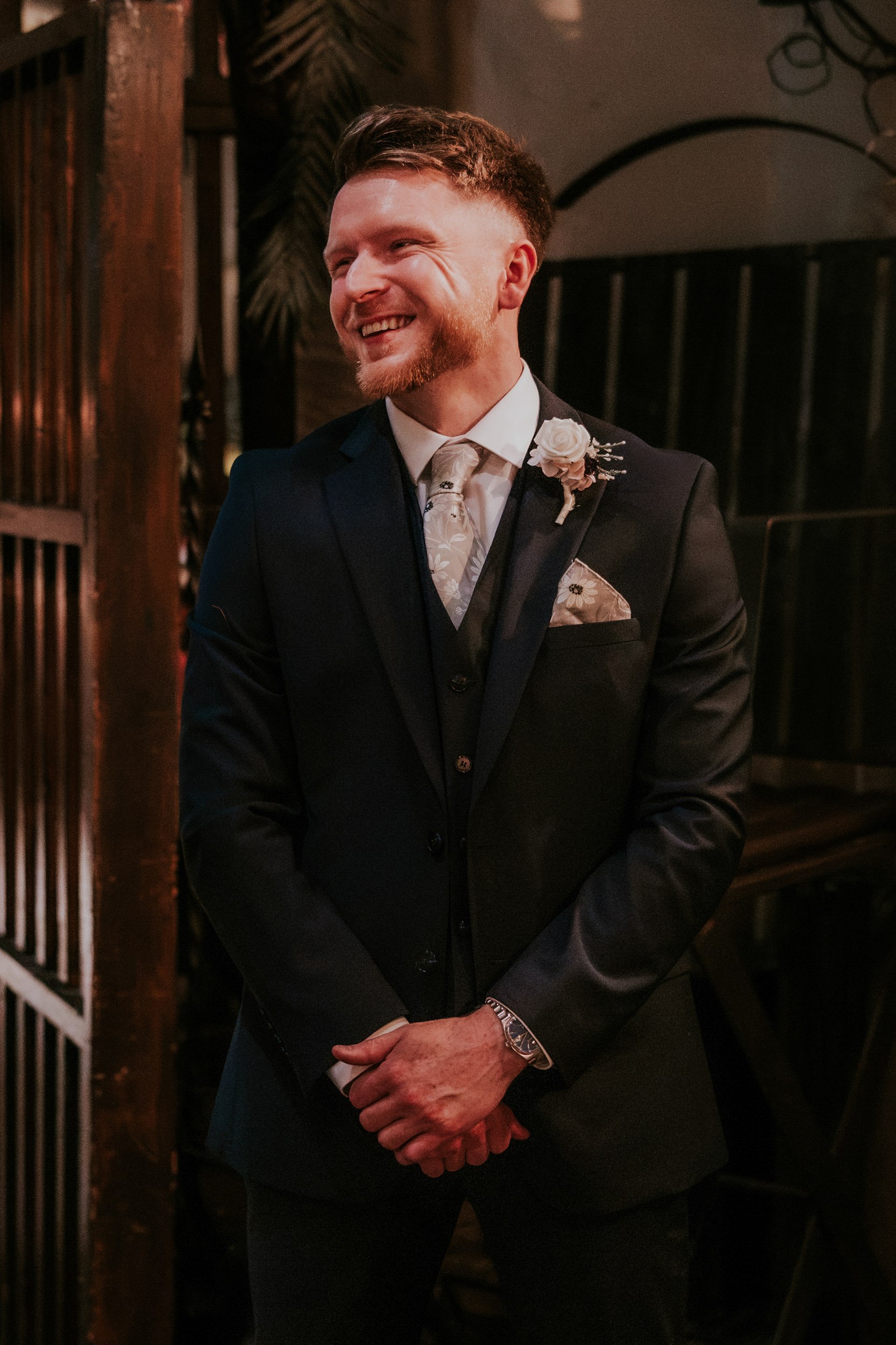 John smiling at Arta in Glasgow, wearing a dark three-piece suit, floral tie and white rose buttonhole.
