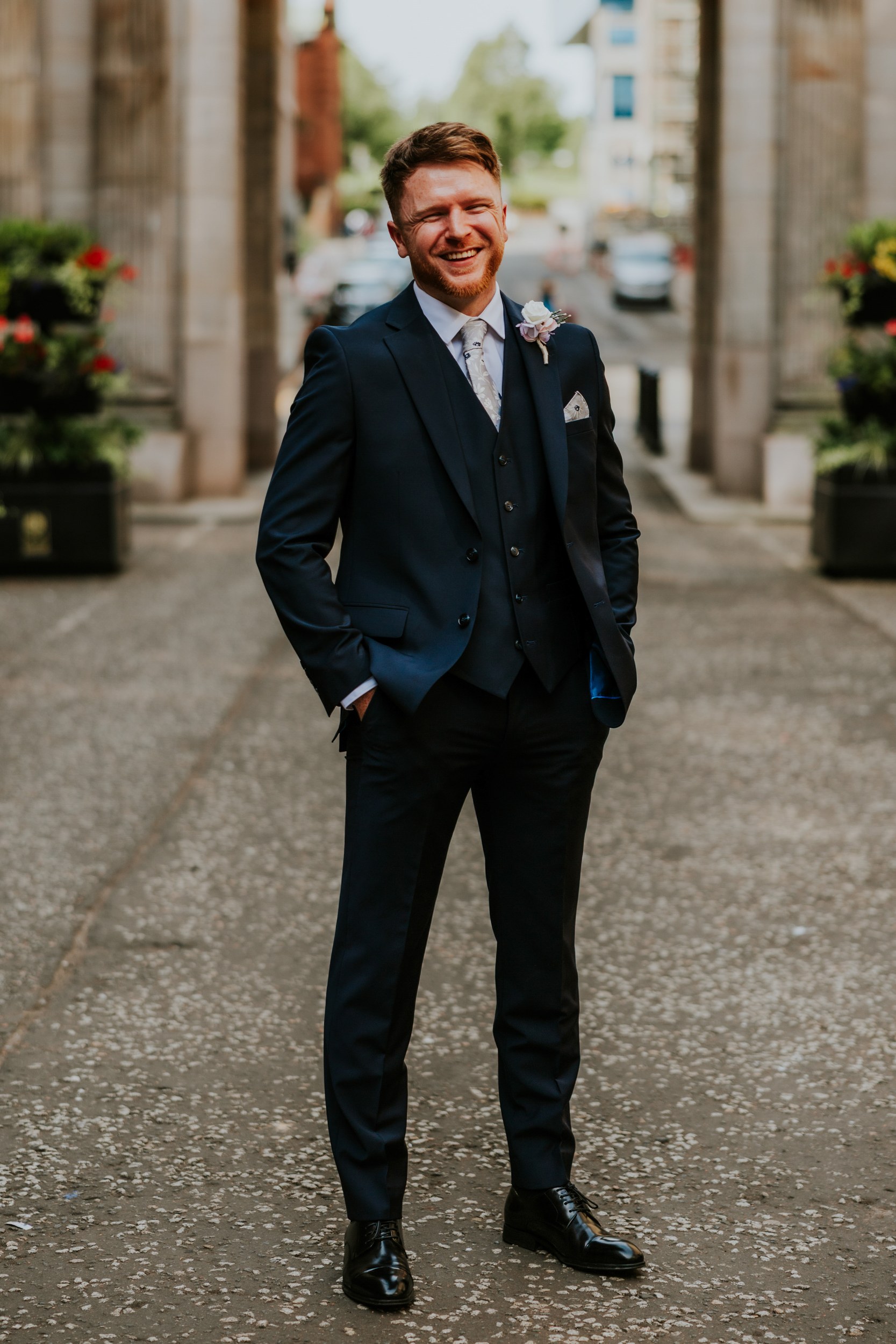 John smiling in a navy three-piece suit with a floral buttonhole, standing between stone columns in Glasgow.