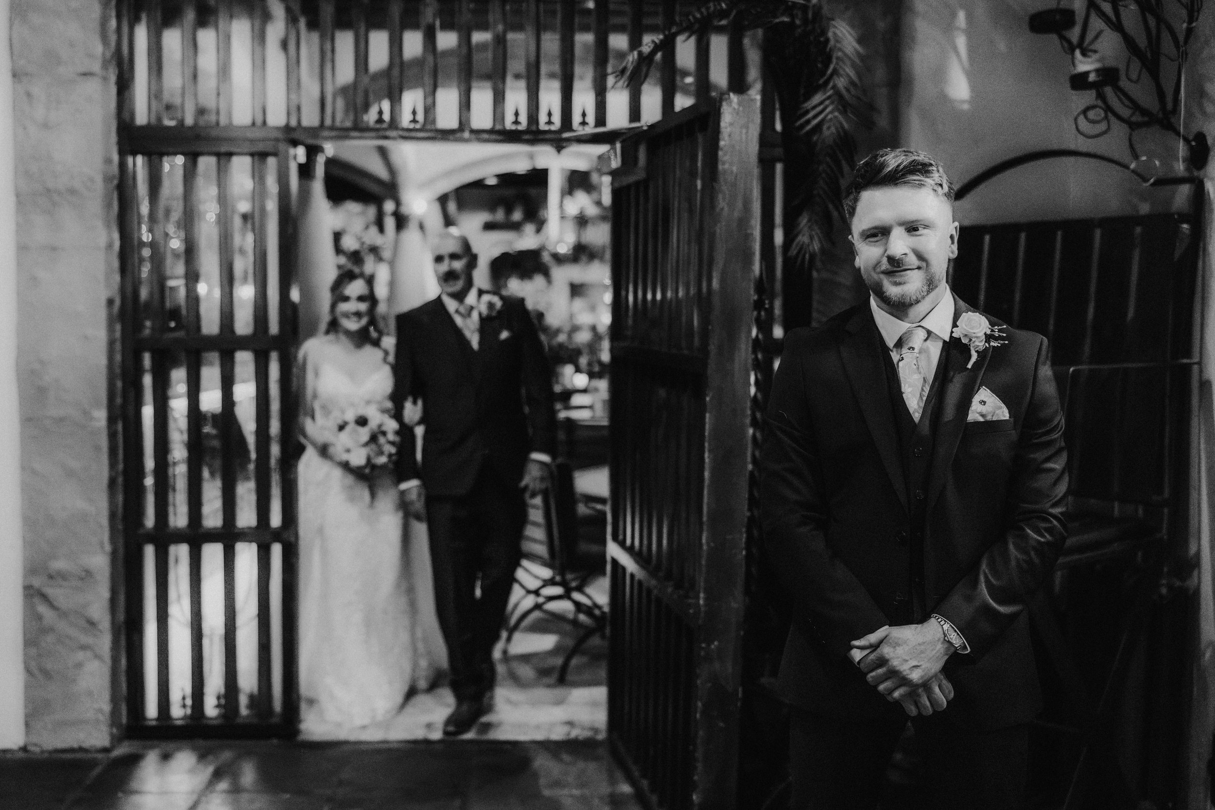 John smiling in his wedding suit at Arta, Glasgow, with Lucy and another man behind him in black and white.