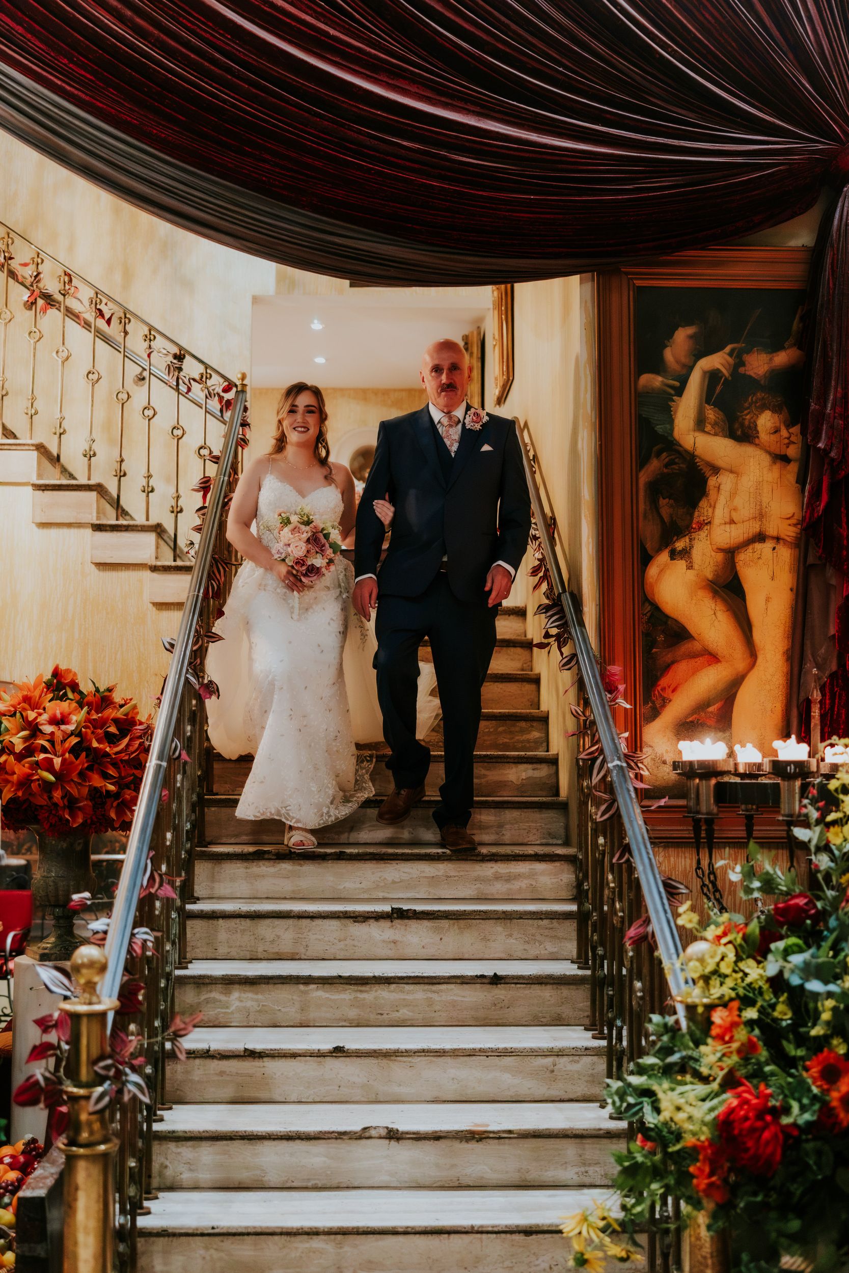 Lucy and John arm in arm on the staircase at Arta, Glasgow, beneath burgundy drapery, with flowers and candles.