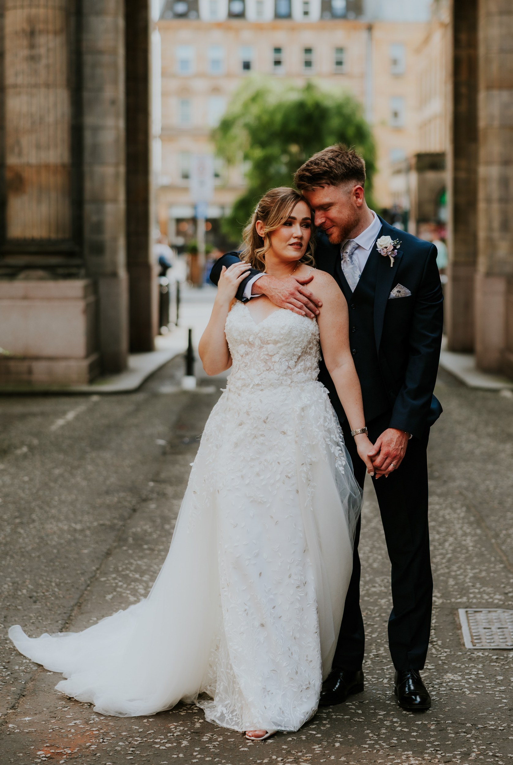 Lucy and John holding hands and embracing between stone columns in Glasgow, wearing their wedding outfits.