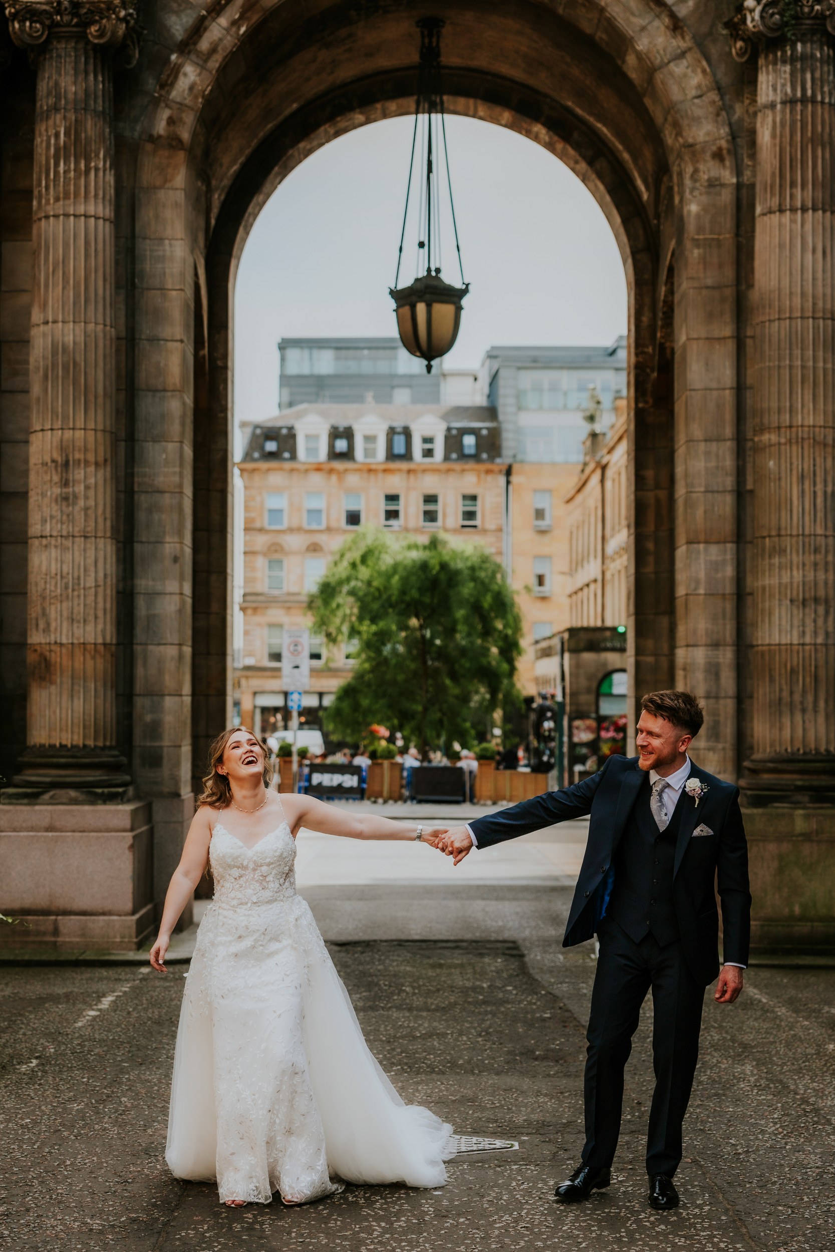 Lucy and John holding hands and laughing beneath a stone archway and hanging lantern in Glasgow.
