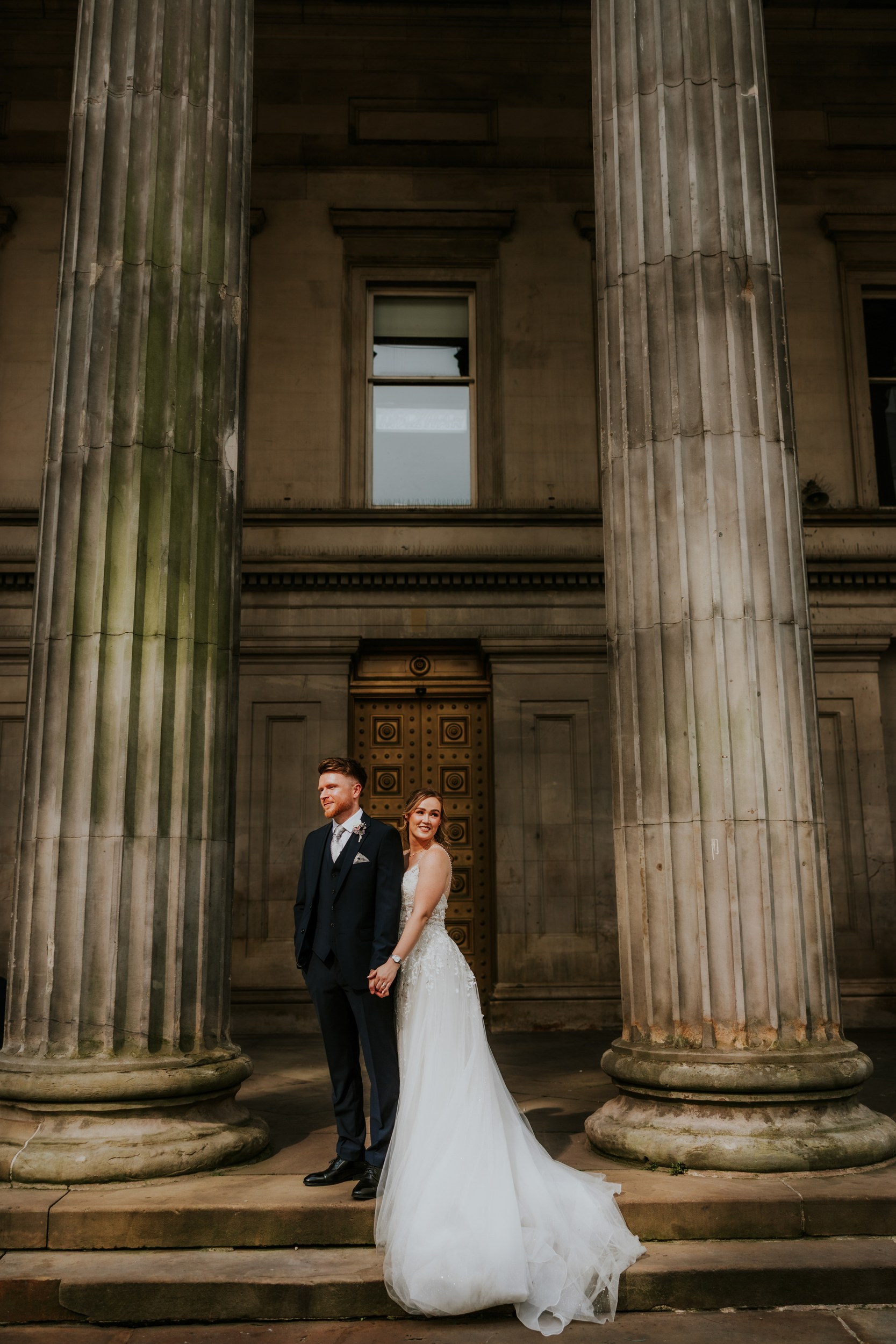 Lucy and John holding hands between tall stone columns in Glasgow, with Lucy’s wedding dress train spread over the steps.