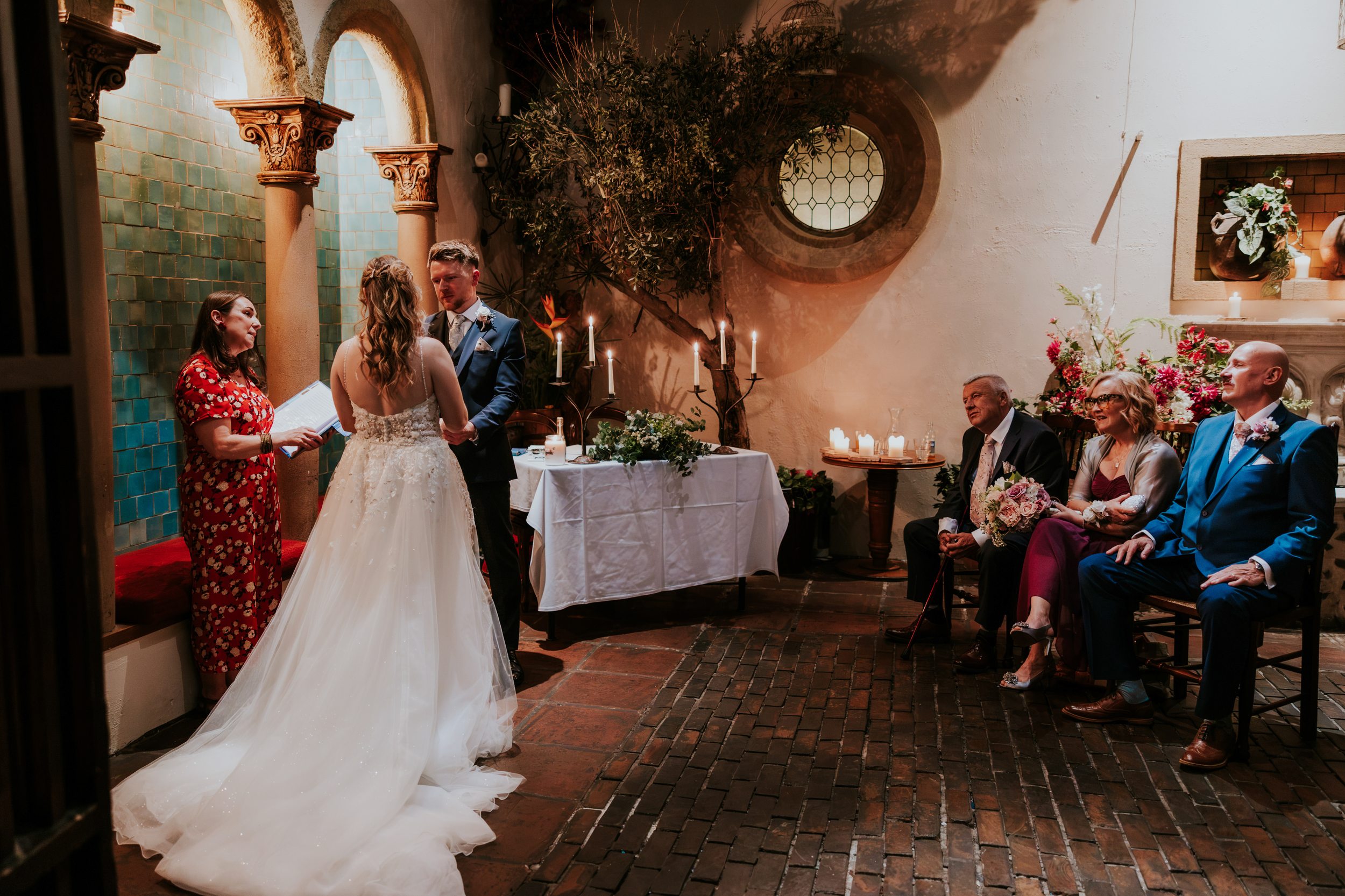 Lucy and John holding hands during their wedding ceremony at Arta, Glasgow, beside their celebrant and seated guests.