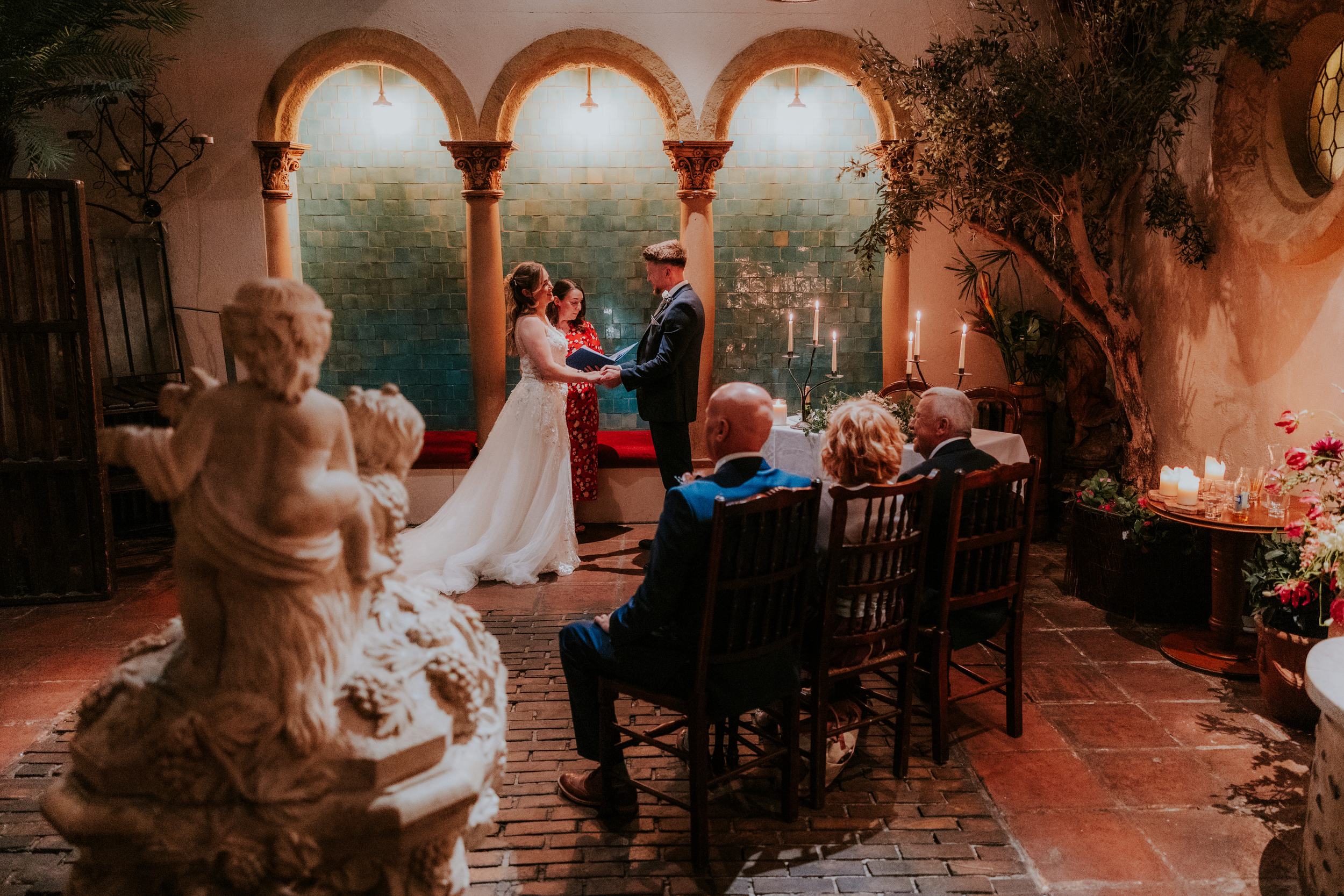Lucy and John holding hands during their wedding ceremony beneath stone arches at Arta, Glasgow.