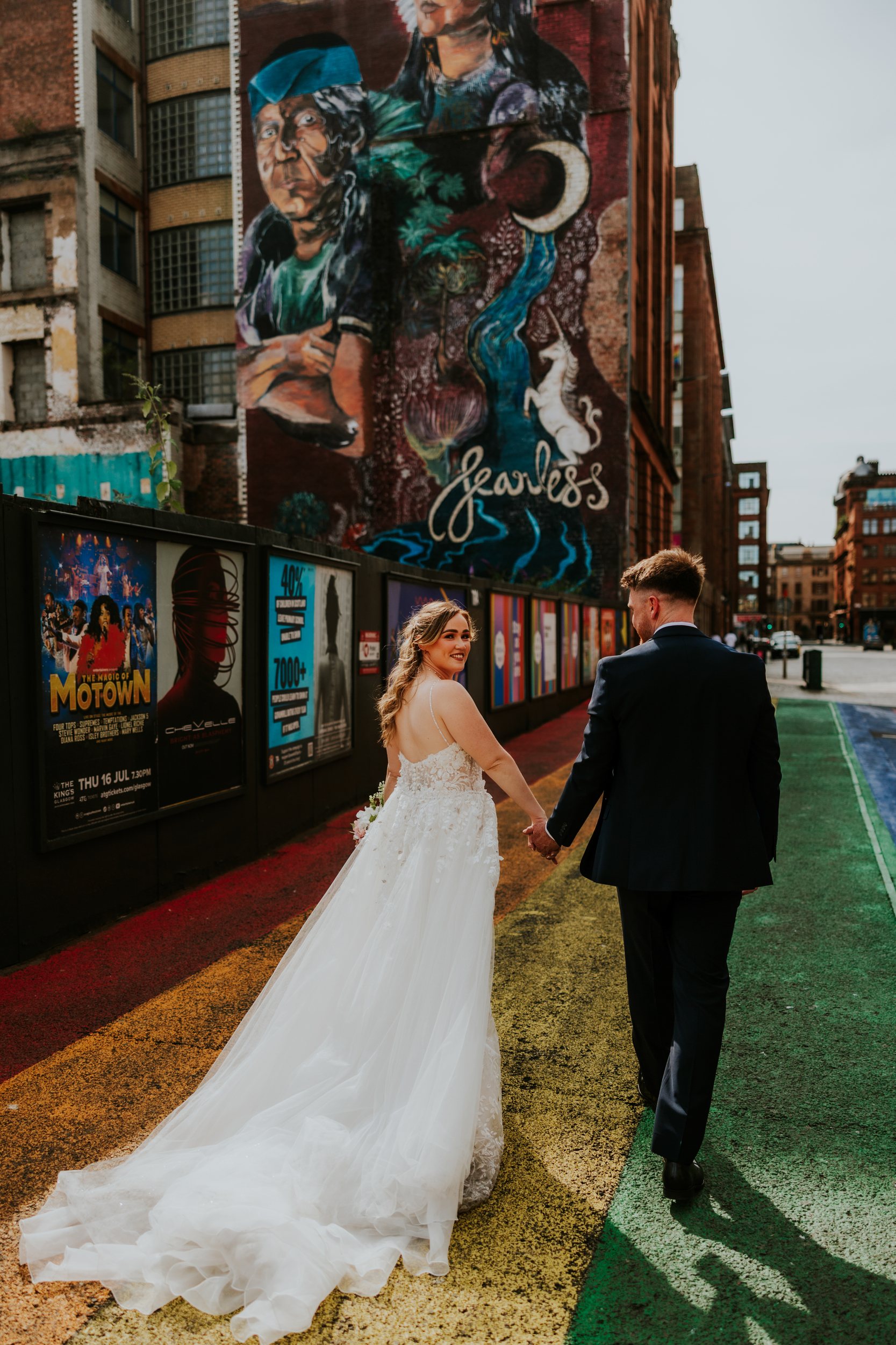 Lucy and John holding hands on a rainbow-painted Glasgow street beneath a mural, with Lucy smiling back at the camera.