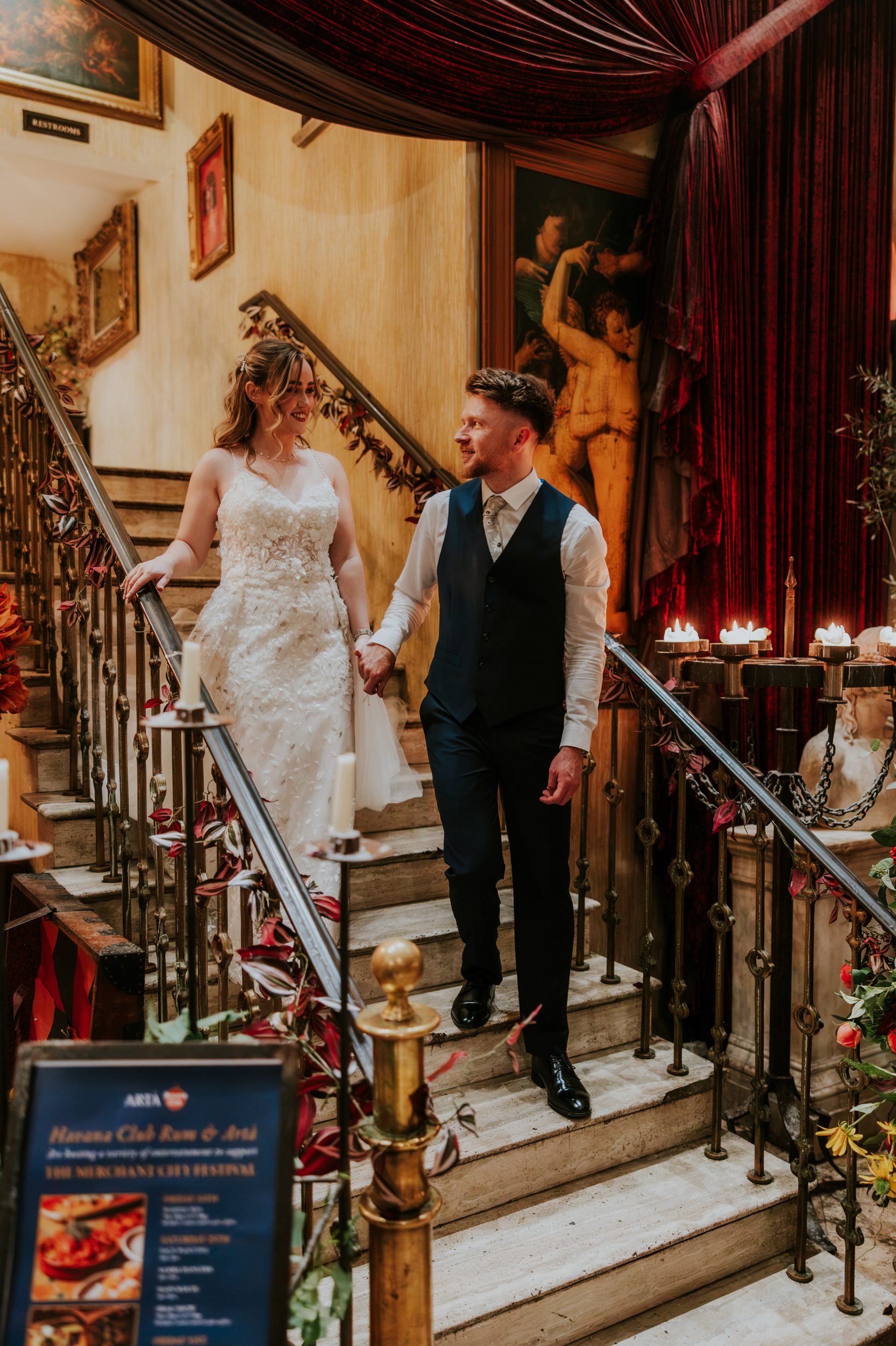 Lucy and John holding hands on the staircase at Arta, Glasgow, with candles and red velvet drapes.