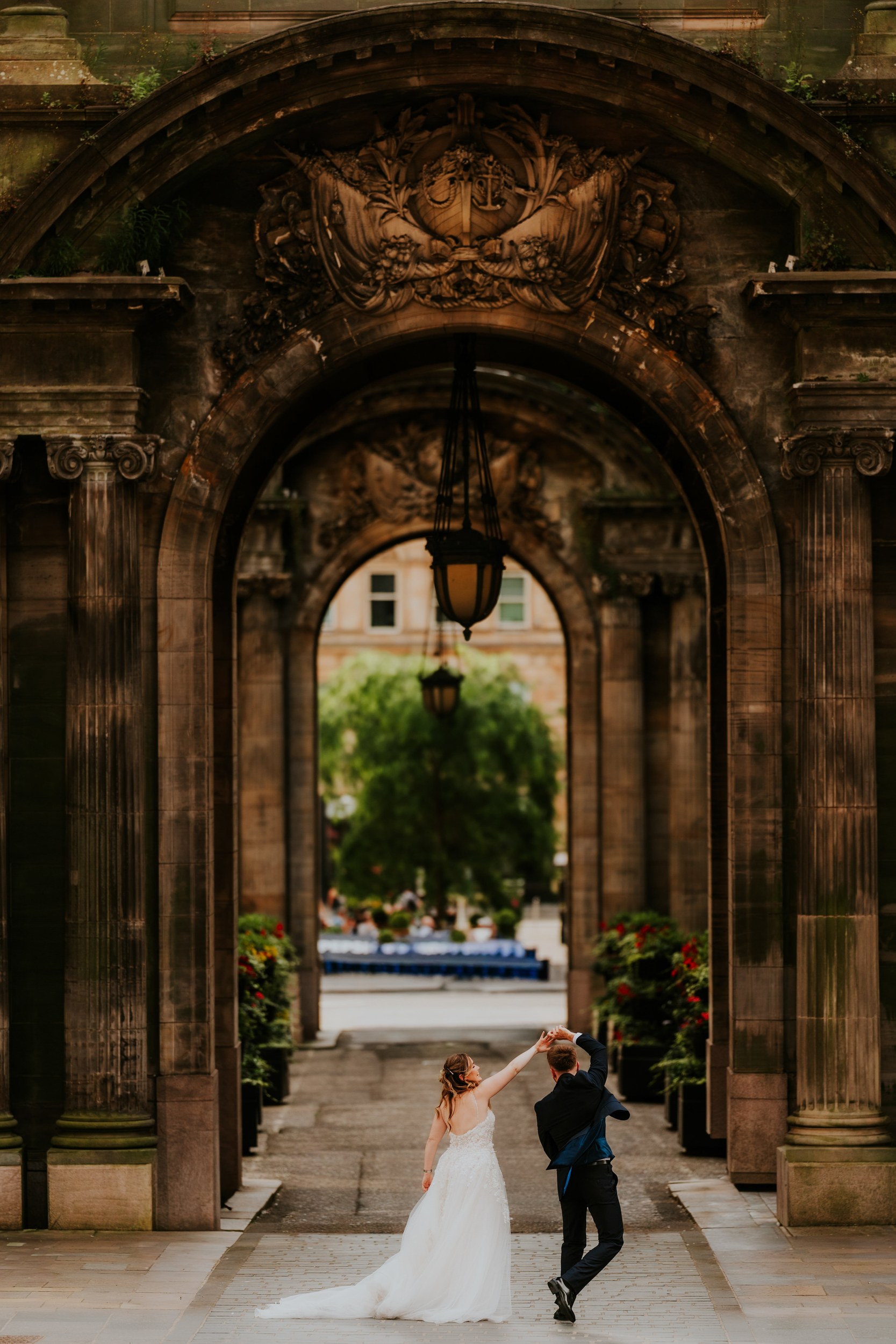 Lucy and John holding hands overhead beneath ornate stone arches and hanging lanterns in Glasgow.