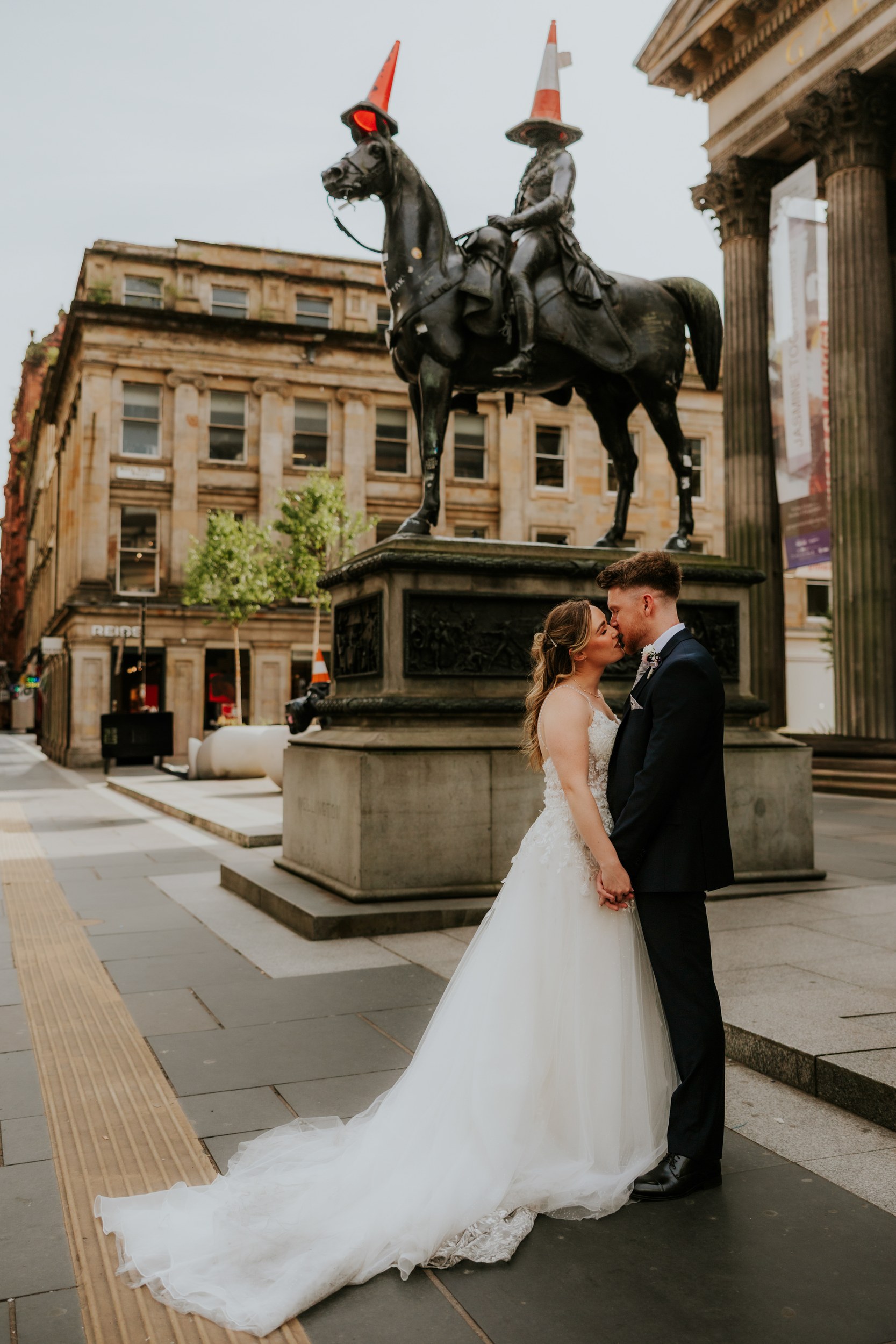 Lucy and John kissing and holding hands beneath Glasgow's Duke of Wellington statue, with traffic cones on the rider and horse.
