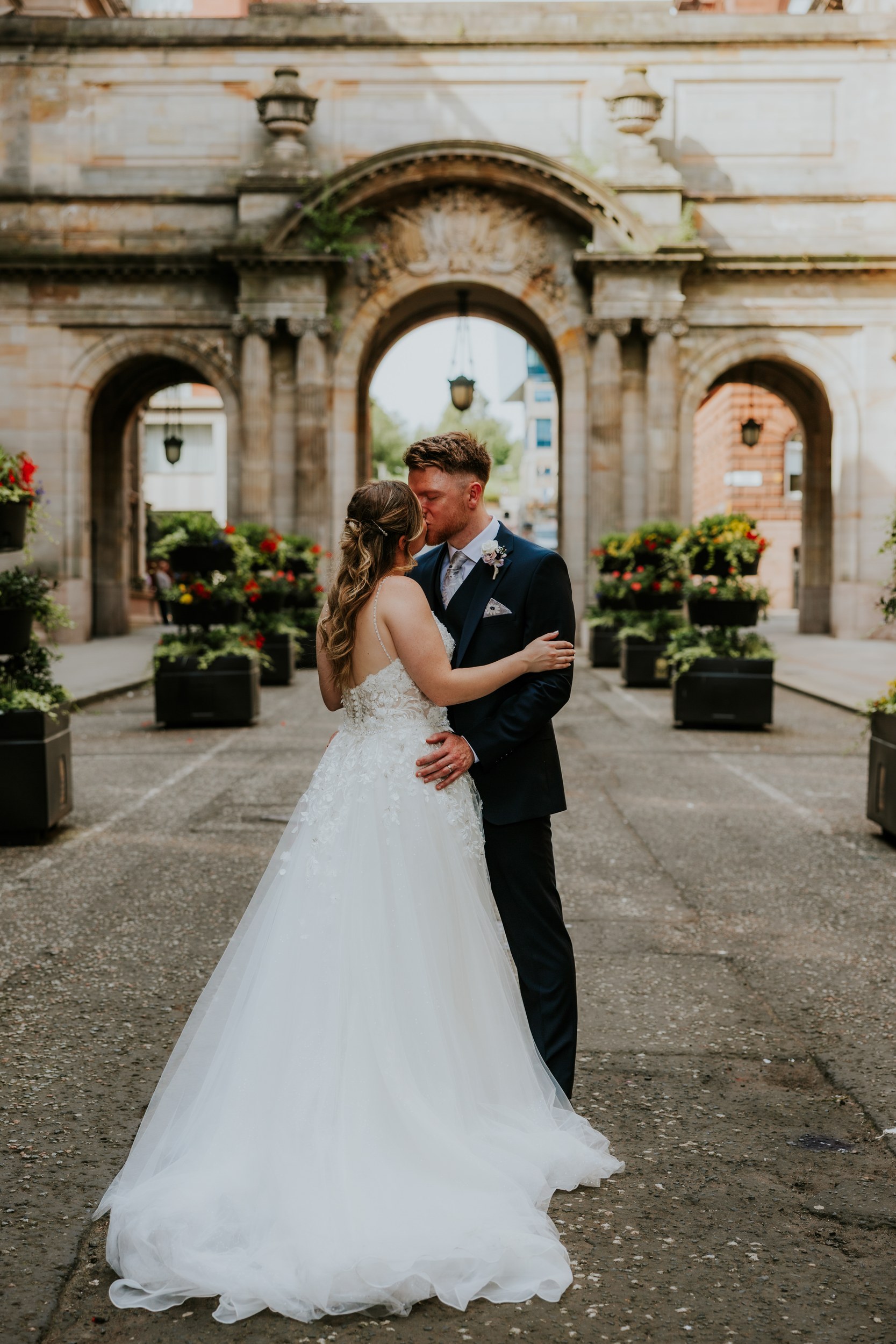 Lucy and John kissing in front of stone arches in Glasgow, with her wedding dress train spread across the paving.