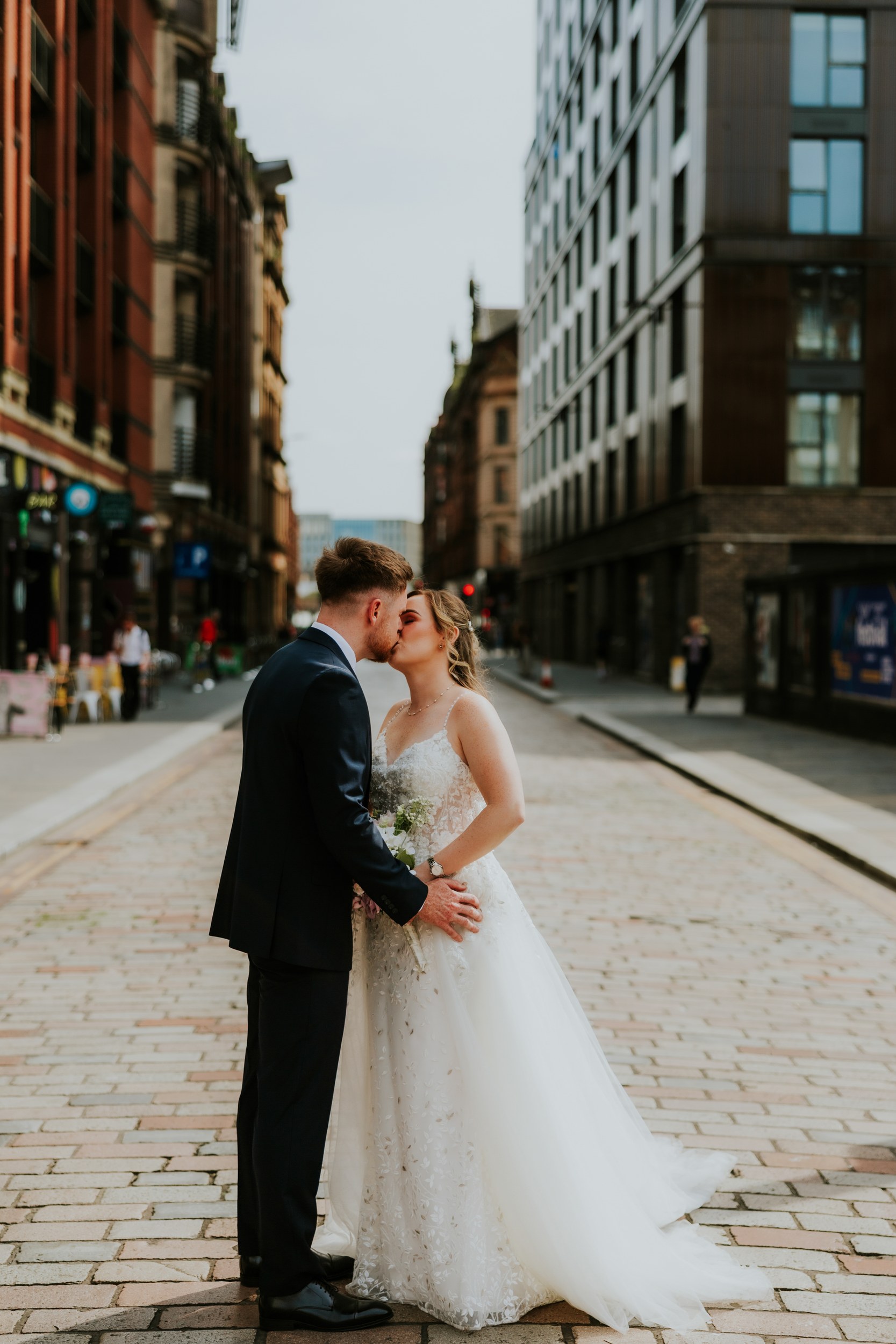 Lucy and John kissing on a cobbled Glasgow street, with Lucy’s lace wedding dress train spread behind her.