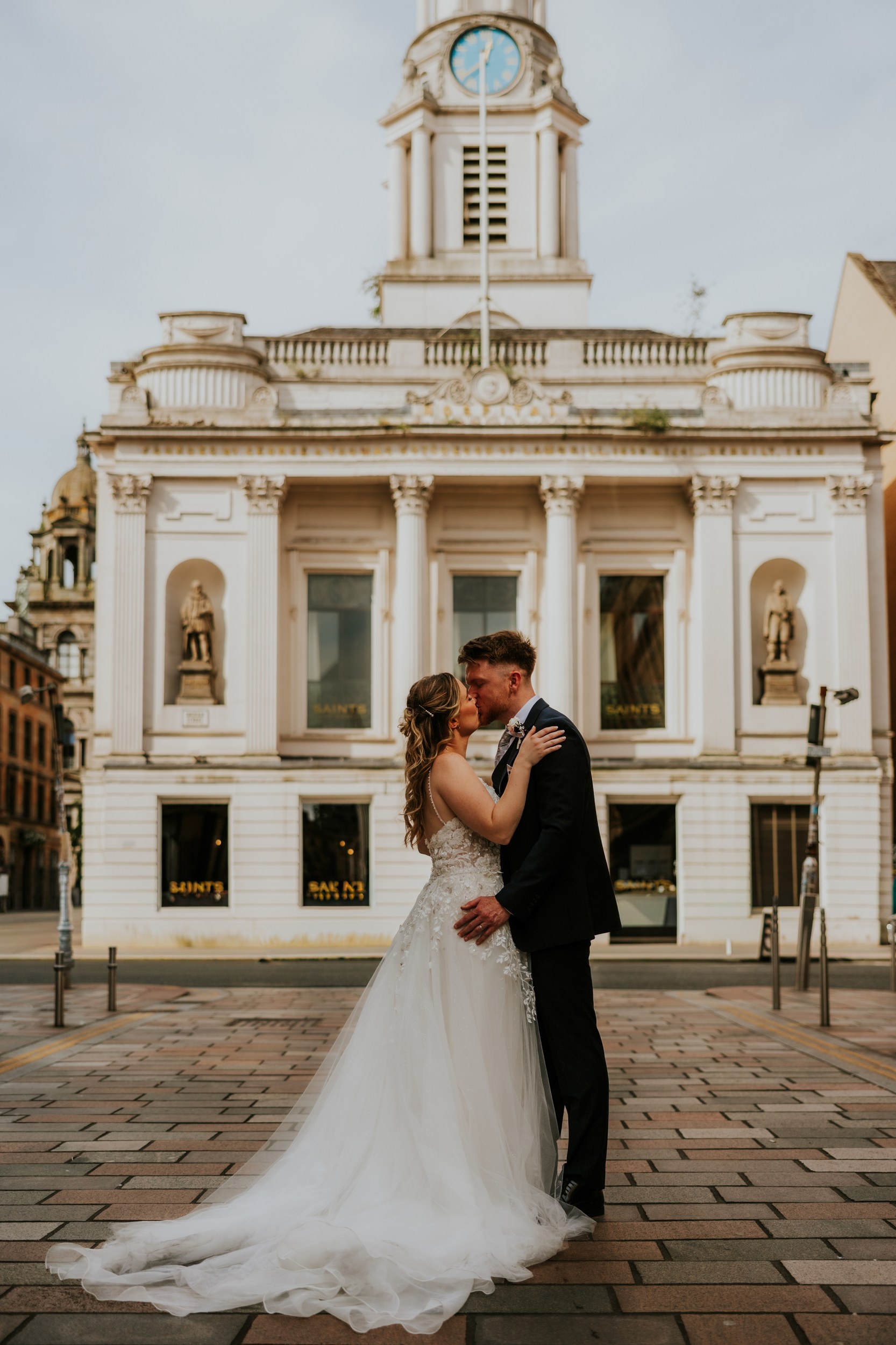 Lucy and John kissing on a Glasgow street, her wedding dress train spread across the paving beneath a clock tower.