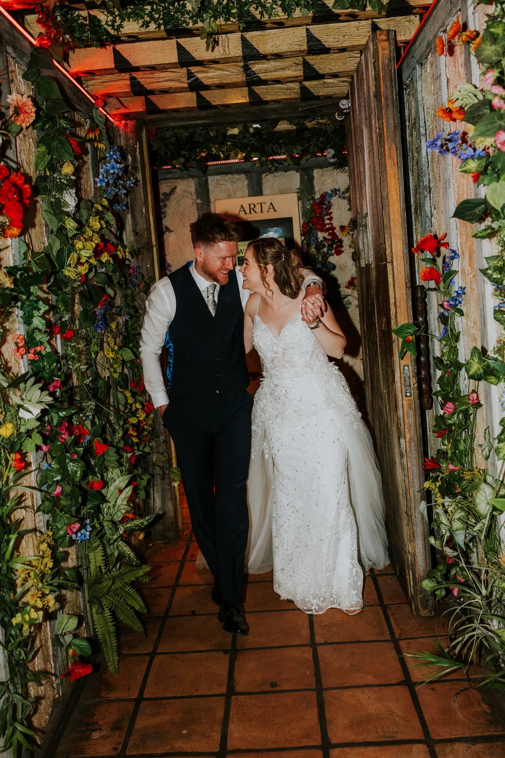 Lucy and John smiling at each other with his arm around her in Arta's flower-lined corridor in Glasgow.