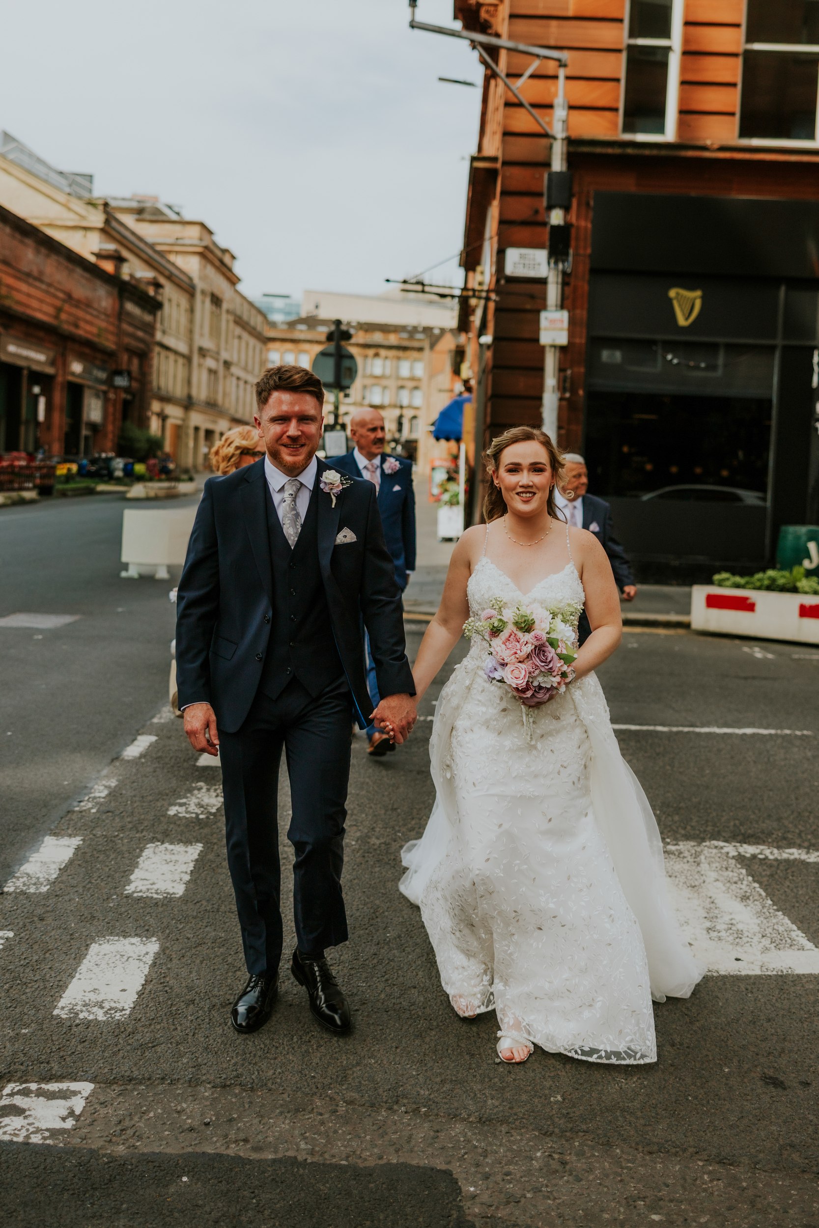 Lucy and John walking hand in hand across a Glasgow street, with Lucy carrying a pastel bouquet and guests following.