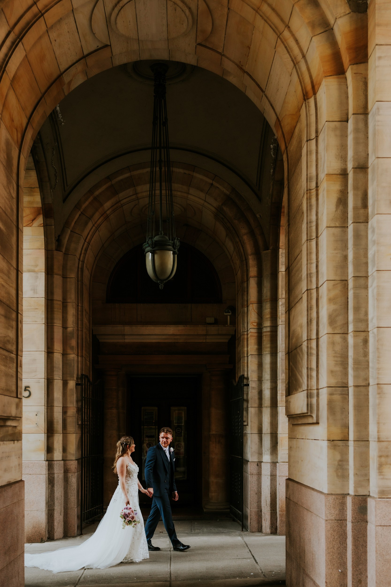 Lucy and John walking hand in hand beneath a stone archway in Glasgow, with Lucy carrying her bouquet.