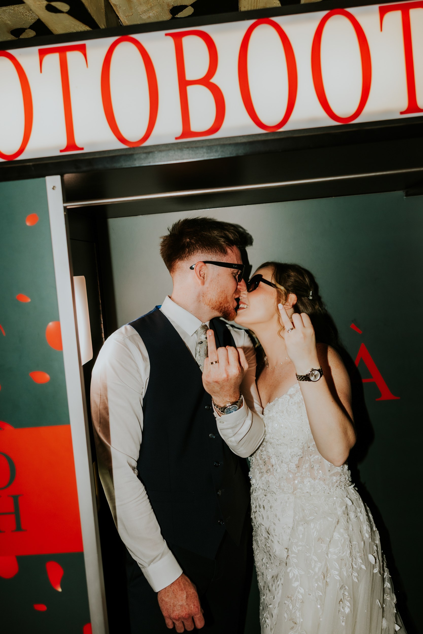 Lucy and John wearing sunglasses in a photo booth, leaning together and holding up their ring fingers.