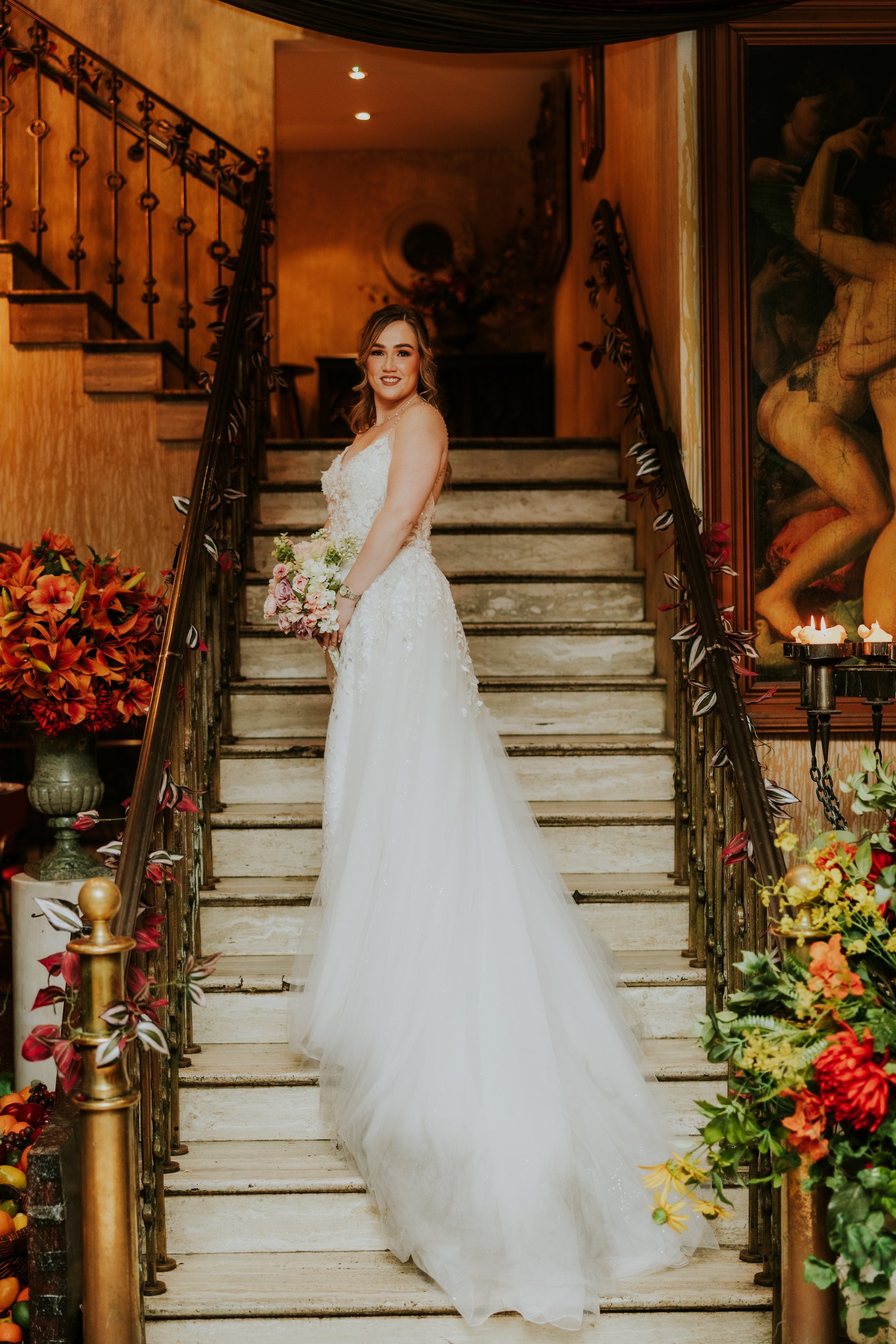 Lucy holding her bouquet on Arta's ornate staircase in Glasgow, with her wedding dress train spread down the steps.
