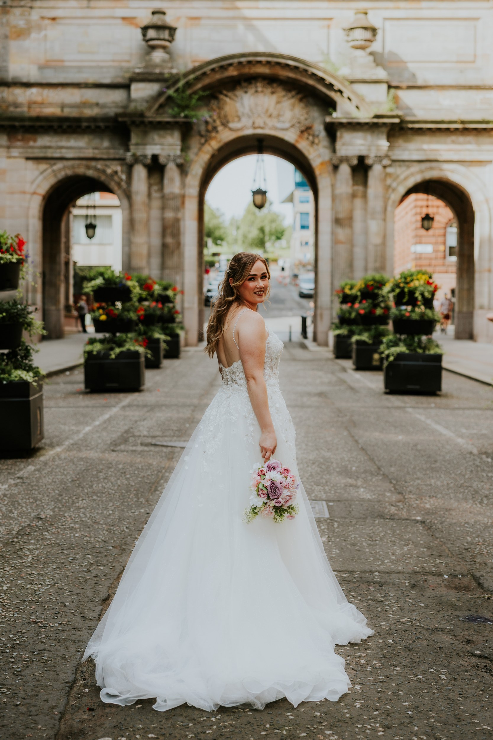 Lucy looking over her shoulder in her wedding dress, holding a pastel bouquet in front of stone arches in Glasgow.