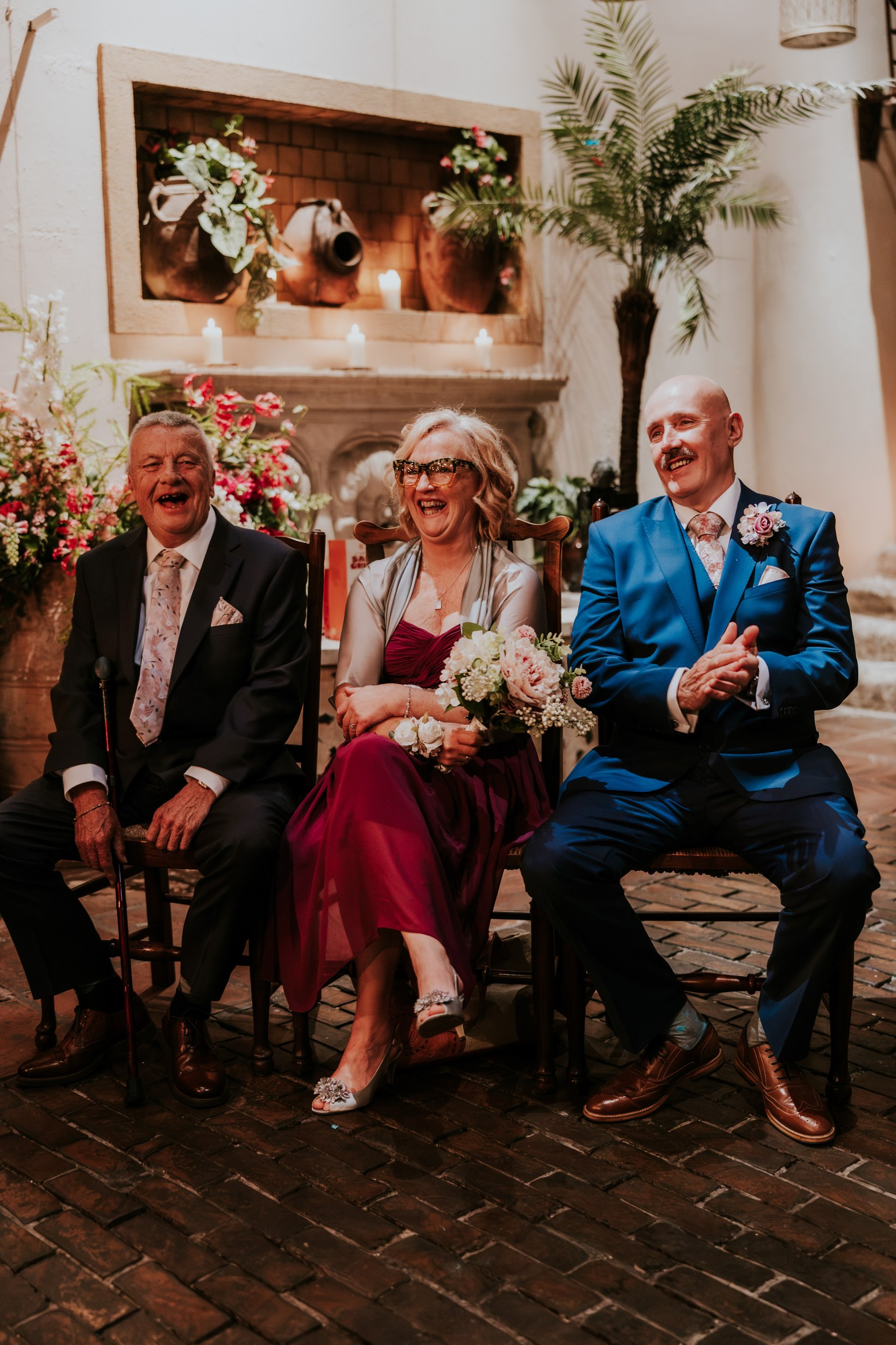 Three people laughing while seated beside flowers and candles at Arta in Glasgow, the woman holding a pale pink bouquet.
