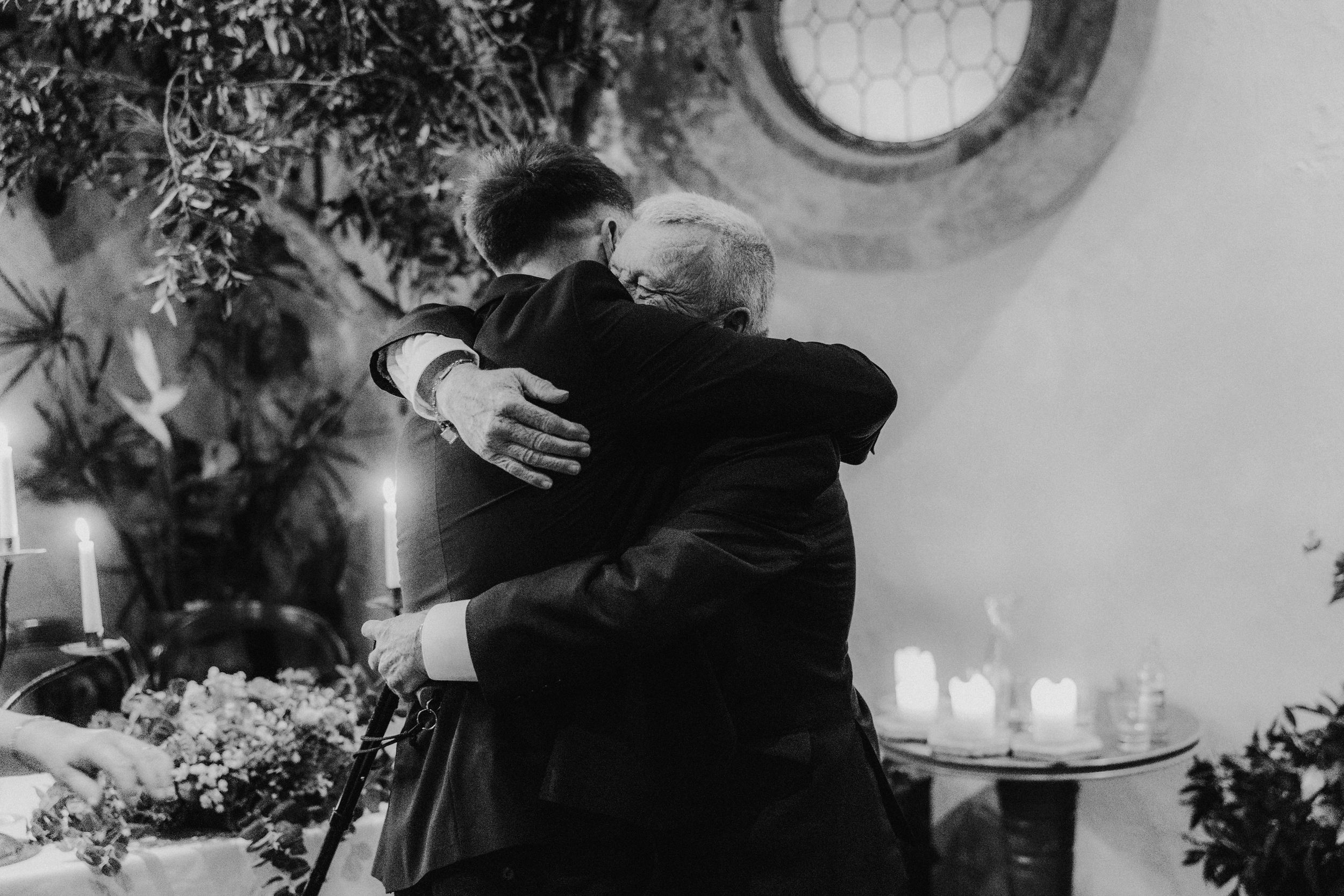 Two men in suits embrace beside lit candles and foliage at Arta in Glasgow, in black and white.