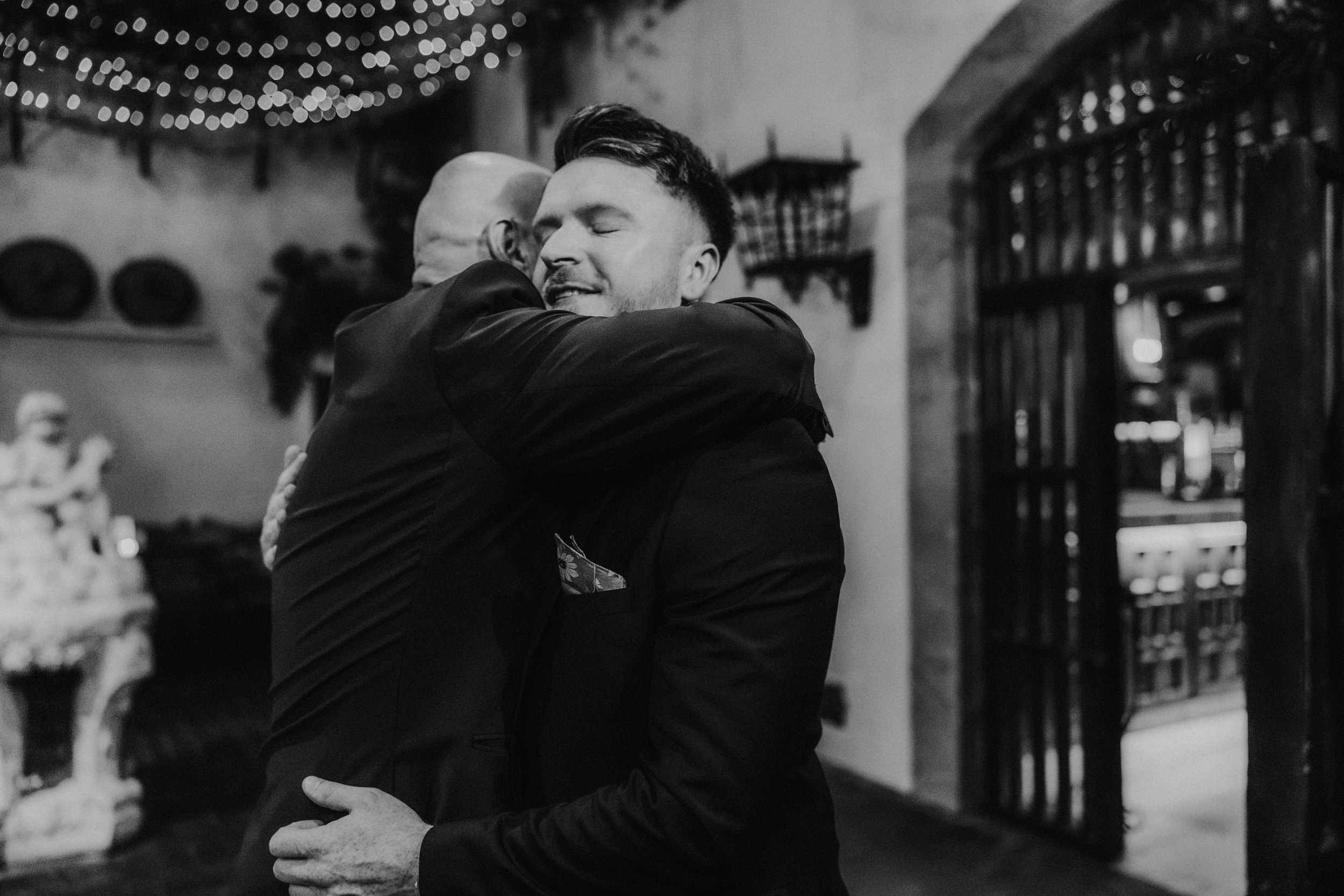 Two men in suits share a hug beneath fairy lights at Arta in Glasgow, in black and white.