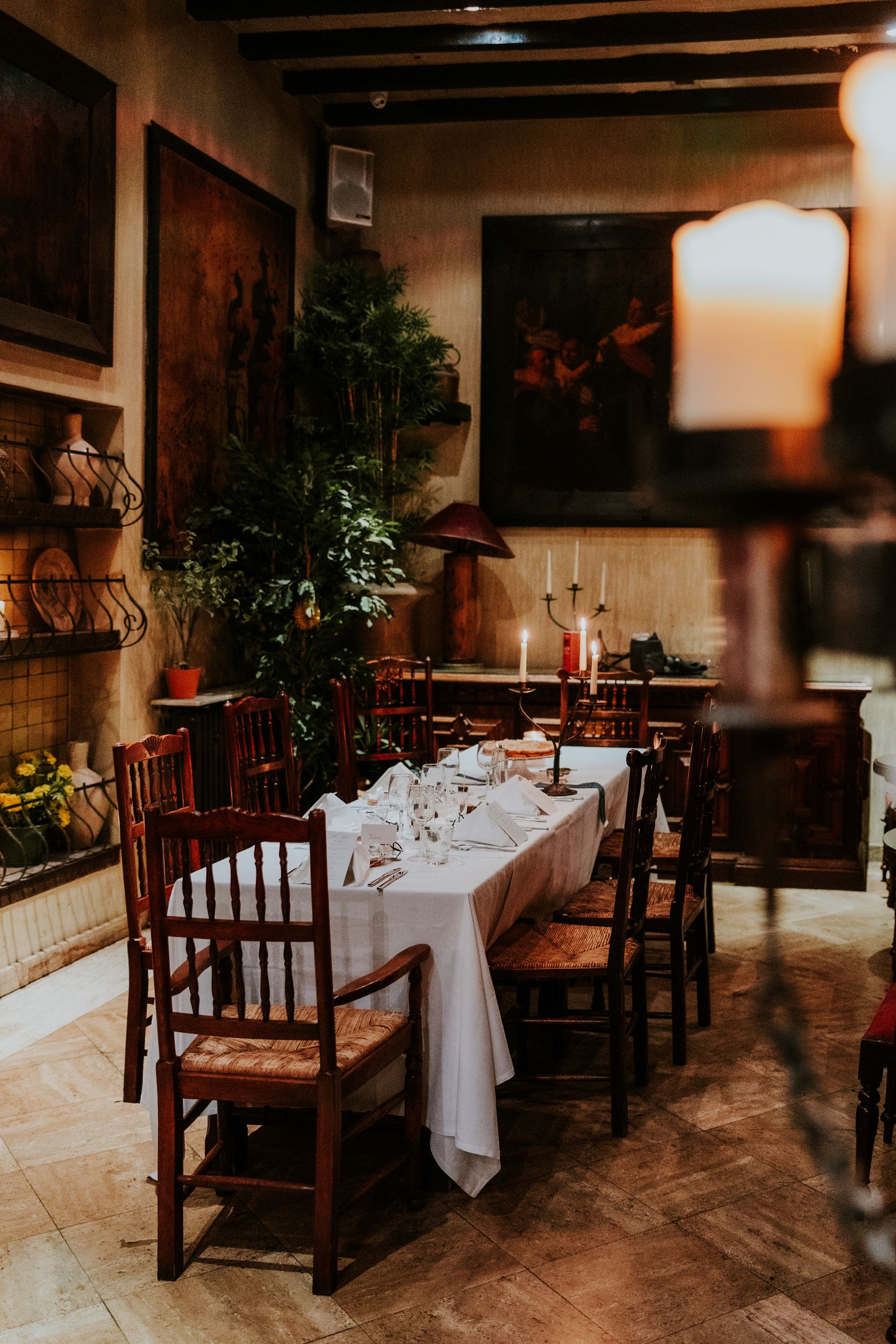 Wedding reception table at Arta in Glasgow, set with white linen, glassware and candles among wooden chairs and plants.