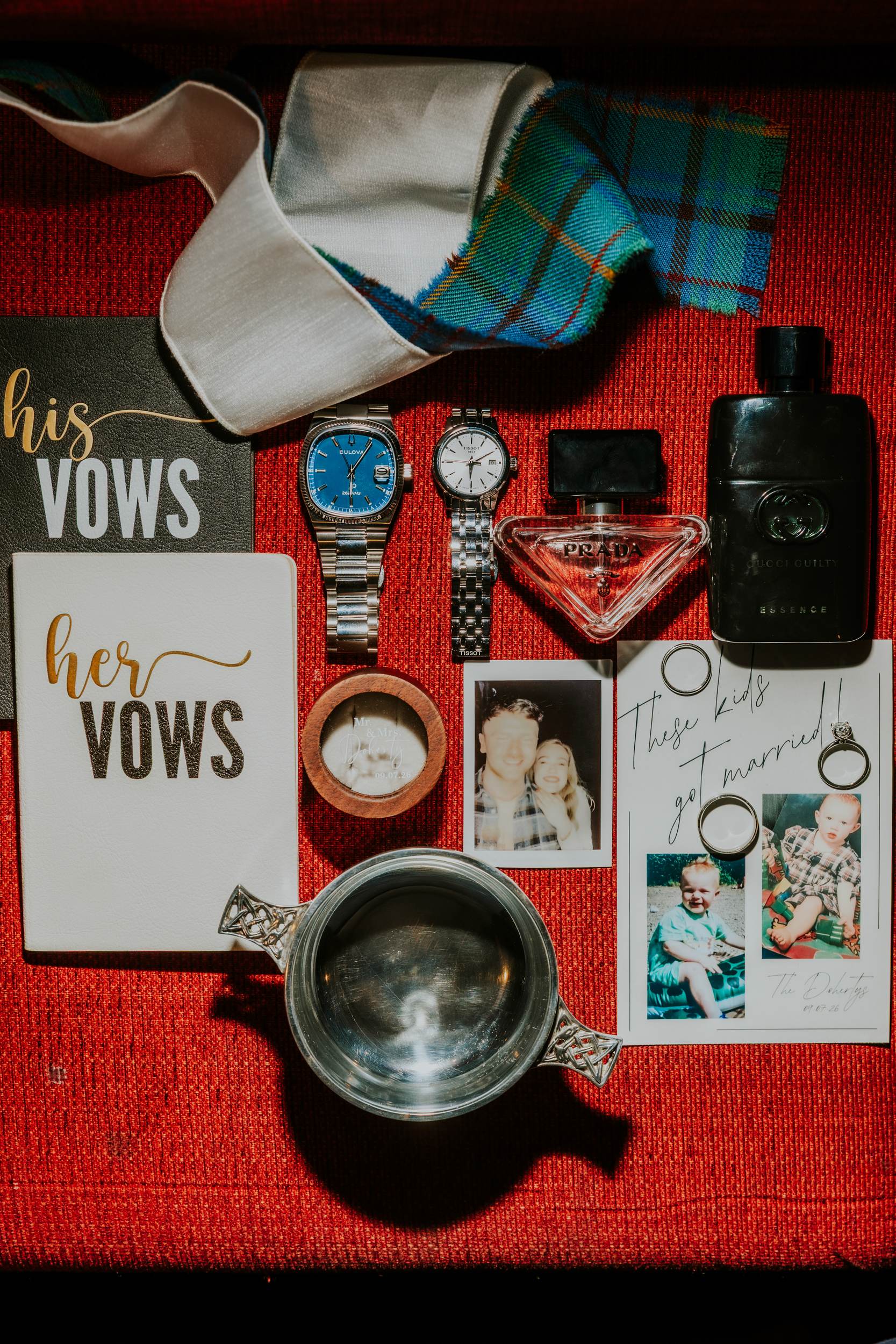 Wedding vow books, rings, watches, fragrances, ribbons and photographs arranged beside a quaich on red fabric.