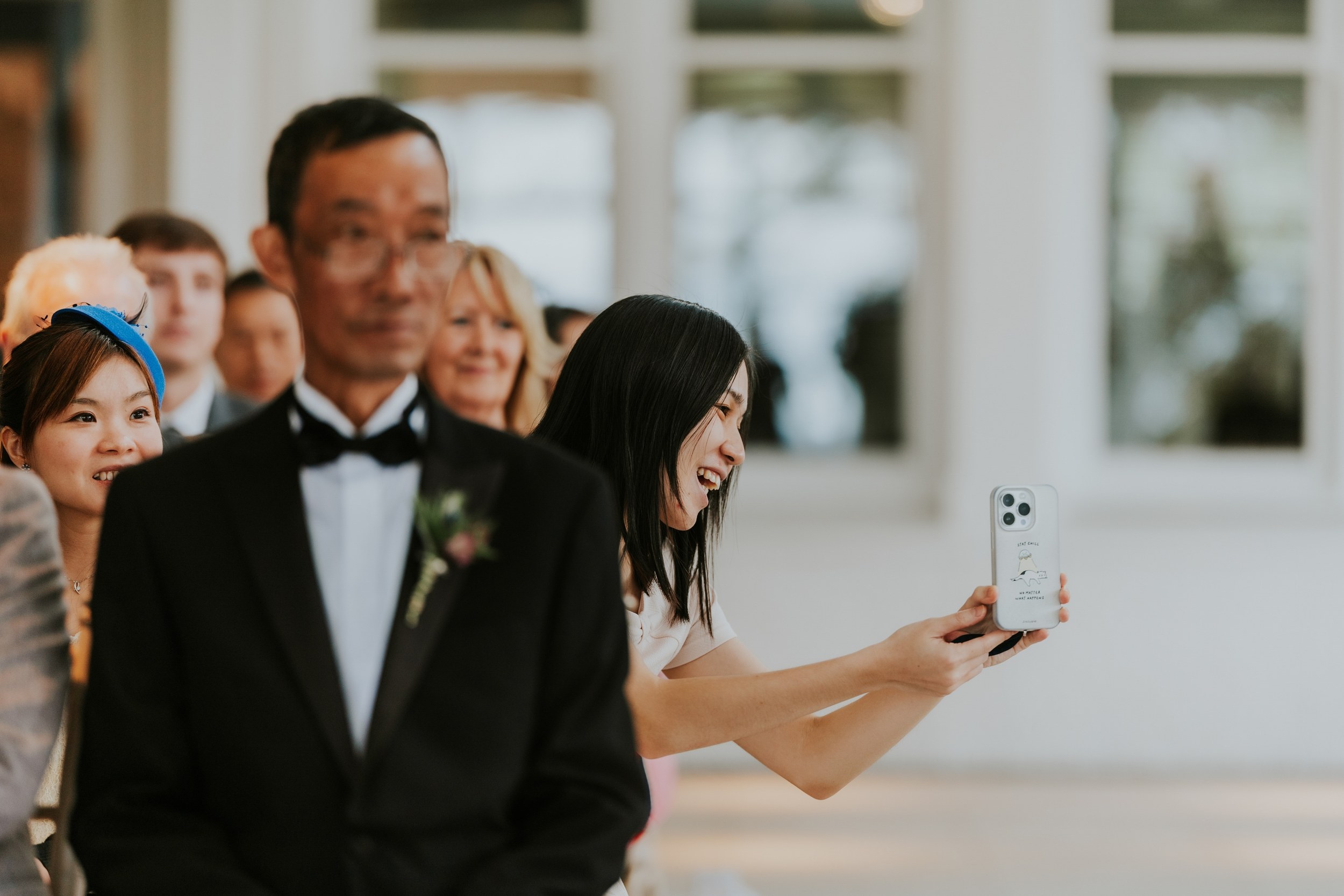 A wedding guest smiles while holding up her phone beside a man in a black dinner jacket at Lochgreen House Hotel.