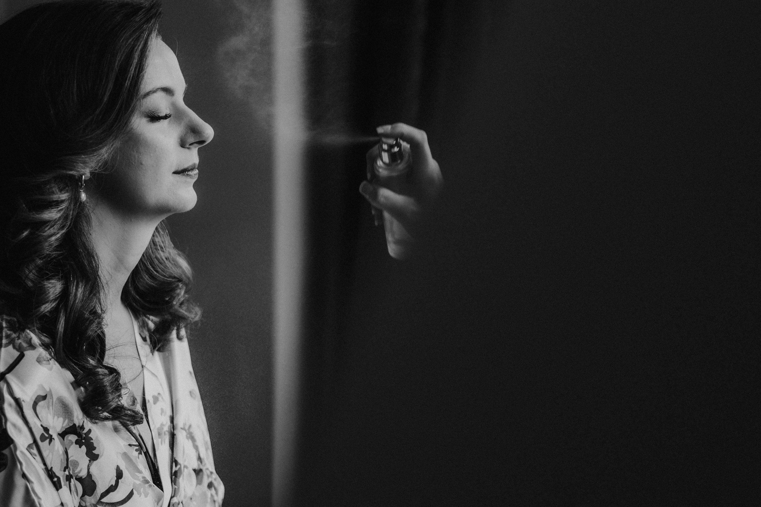 Black-and-white close-up of a woman in a floral robe having her face misted during wedding preparations.