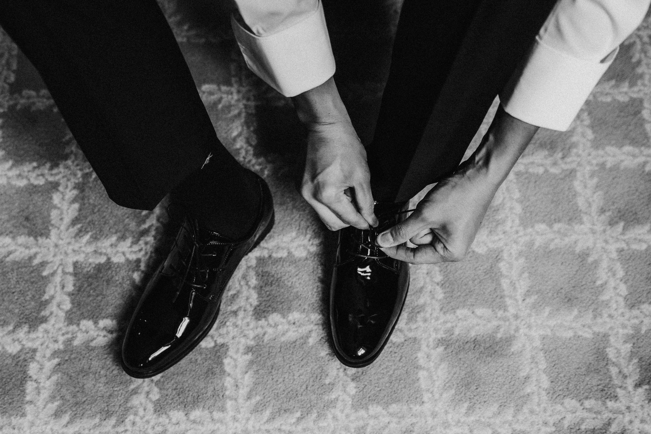 Black-and-white close-up of hands tying the laces of a polished black dress shoe on a patterned carpet.