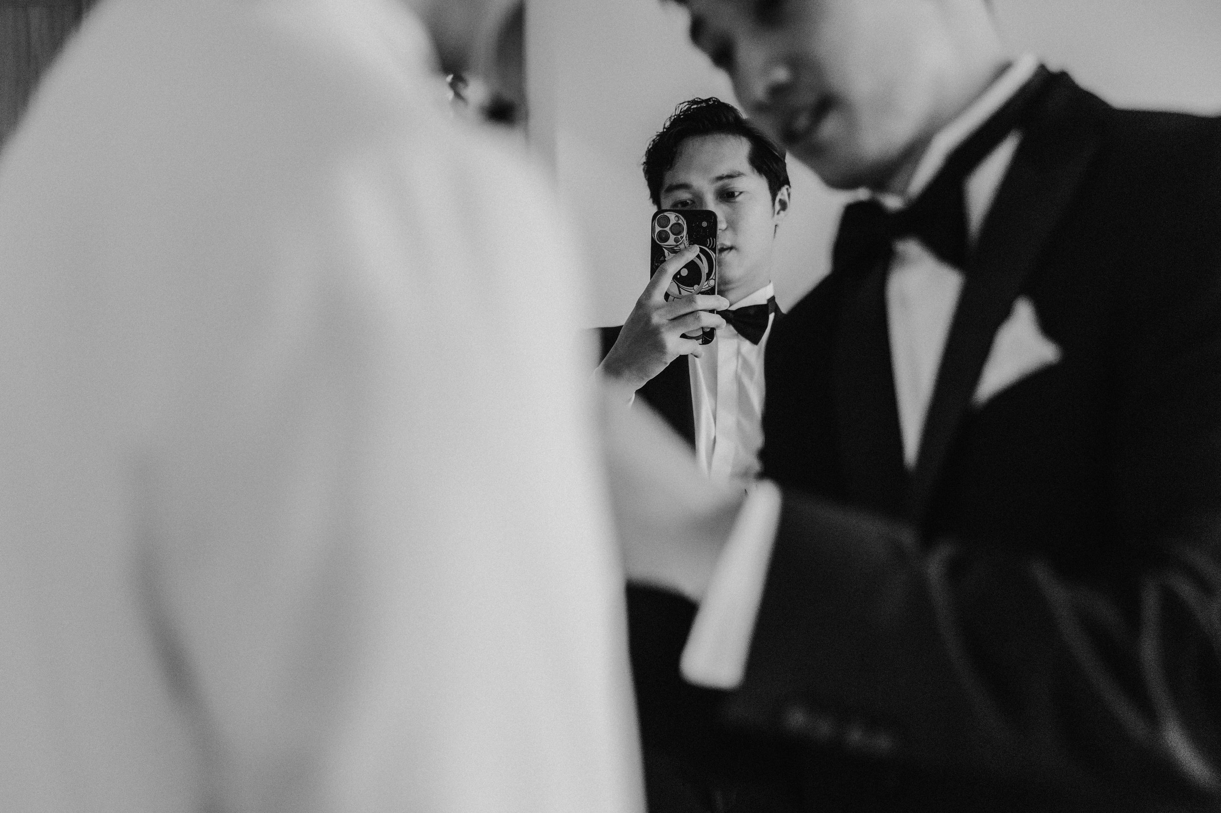 Black-and-white photo of a guest holding up a smartphone between the out-of-focus bride and groom.