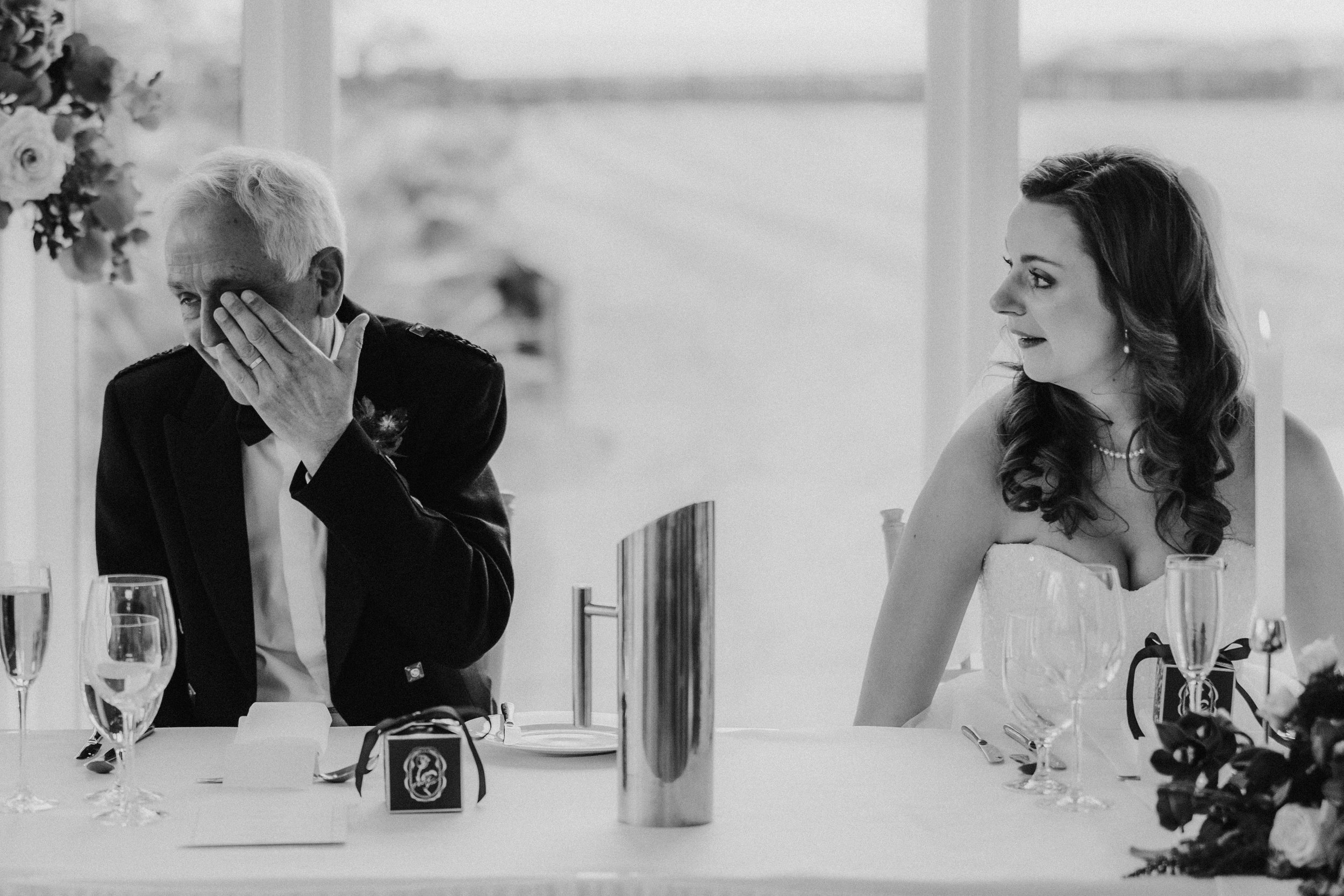 Black-and-white photo of Emma beside a man wiping his eye at the wedding reception table at Lochgreen House Hotel.